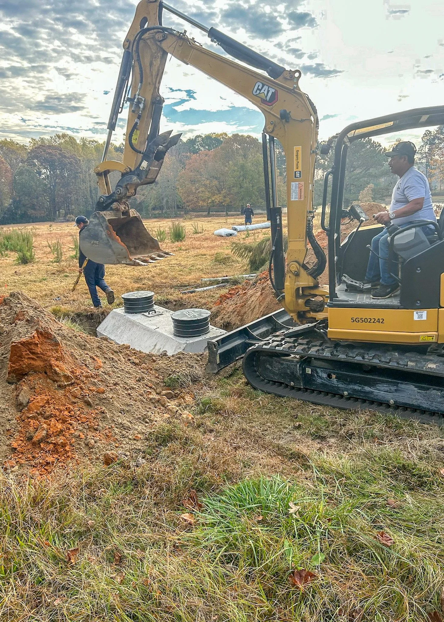 Construction workers are installing underground utility pipes using a yellow Caterpillar excavator on a grassy field with trees in the background.