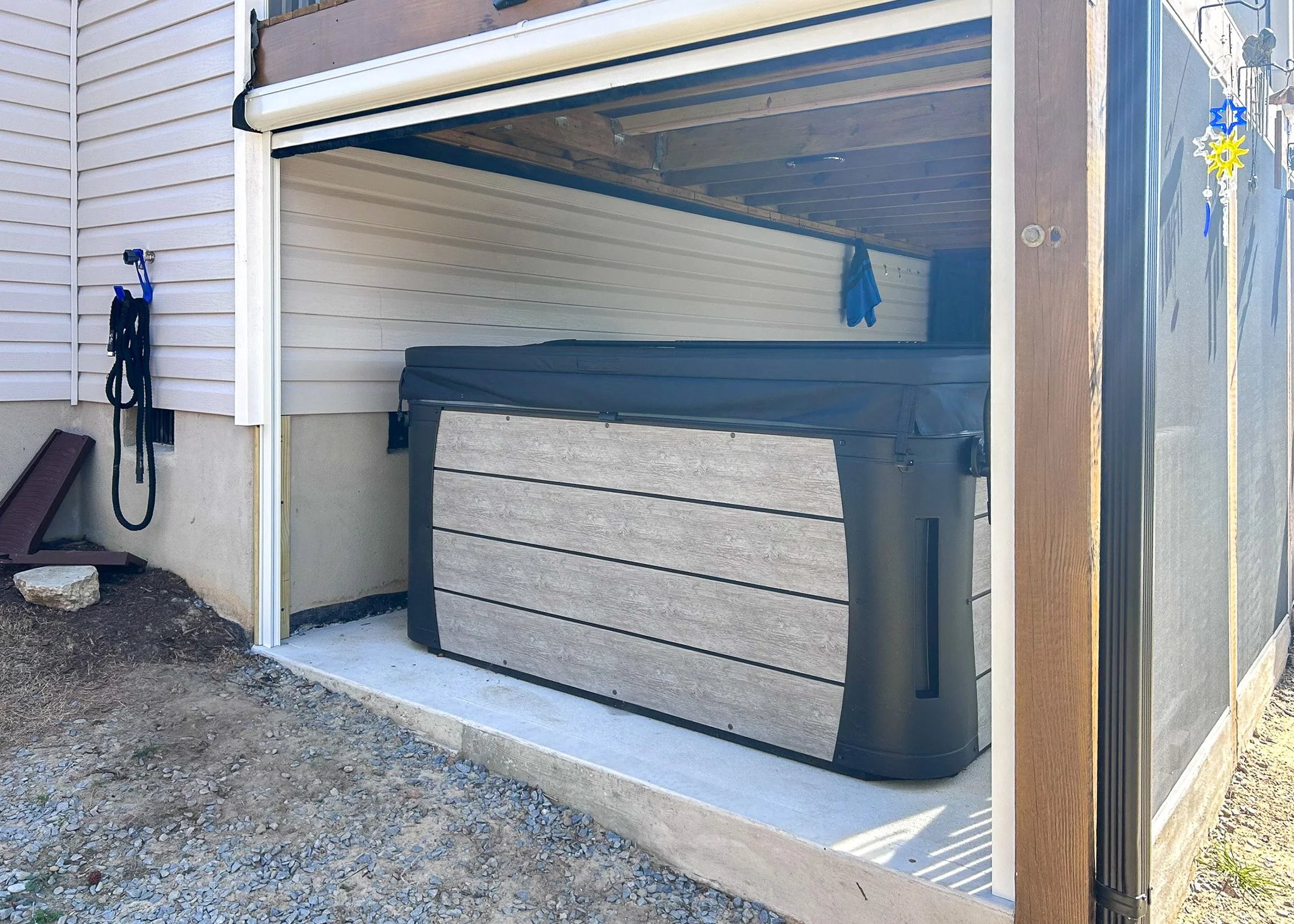A covered outdoor space with a hot tub, a hanging towel, and a black hanging item on a hook on the side of a house with beige siding.