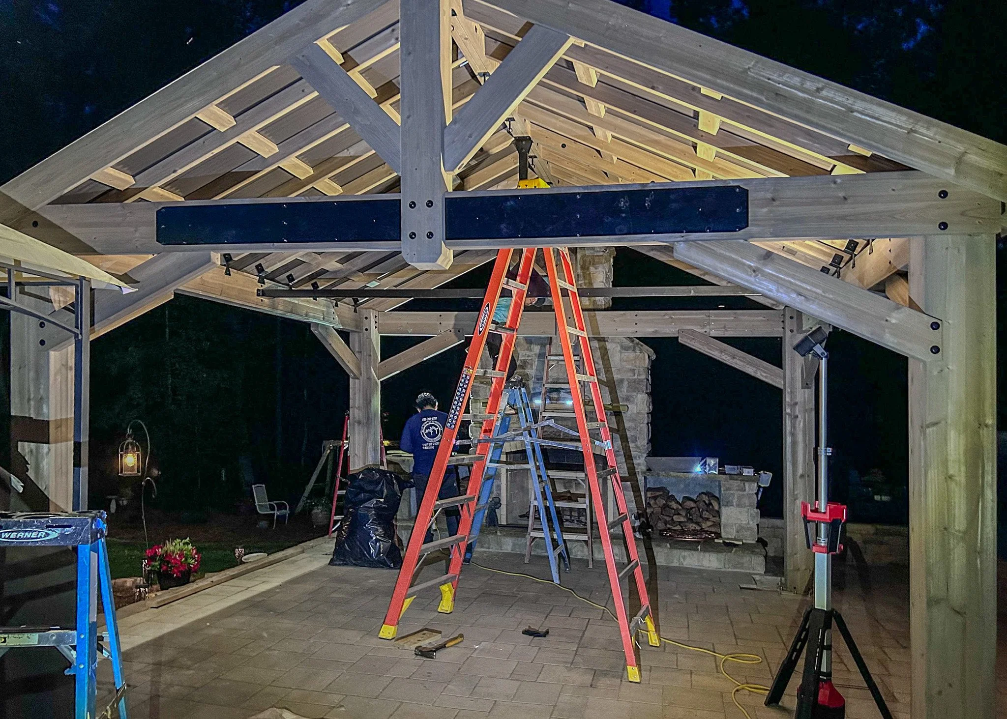 Construction workers building a wooden pergola with a sloped roof at night, using ladders and tools.