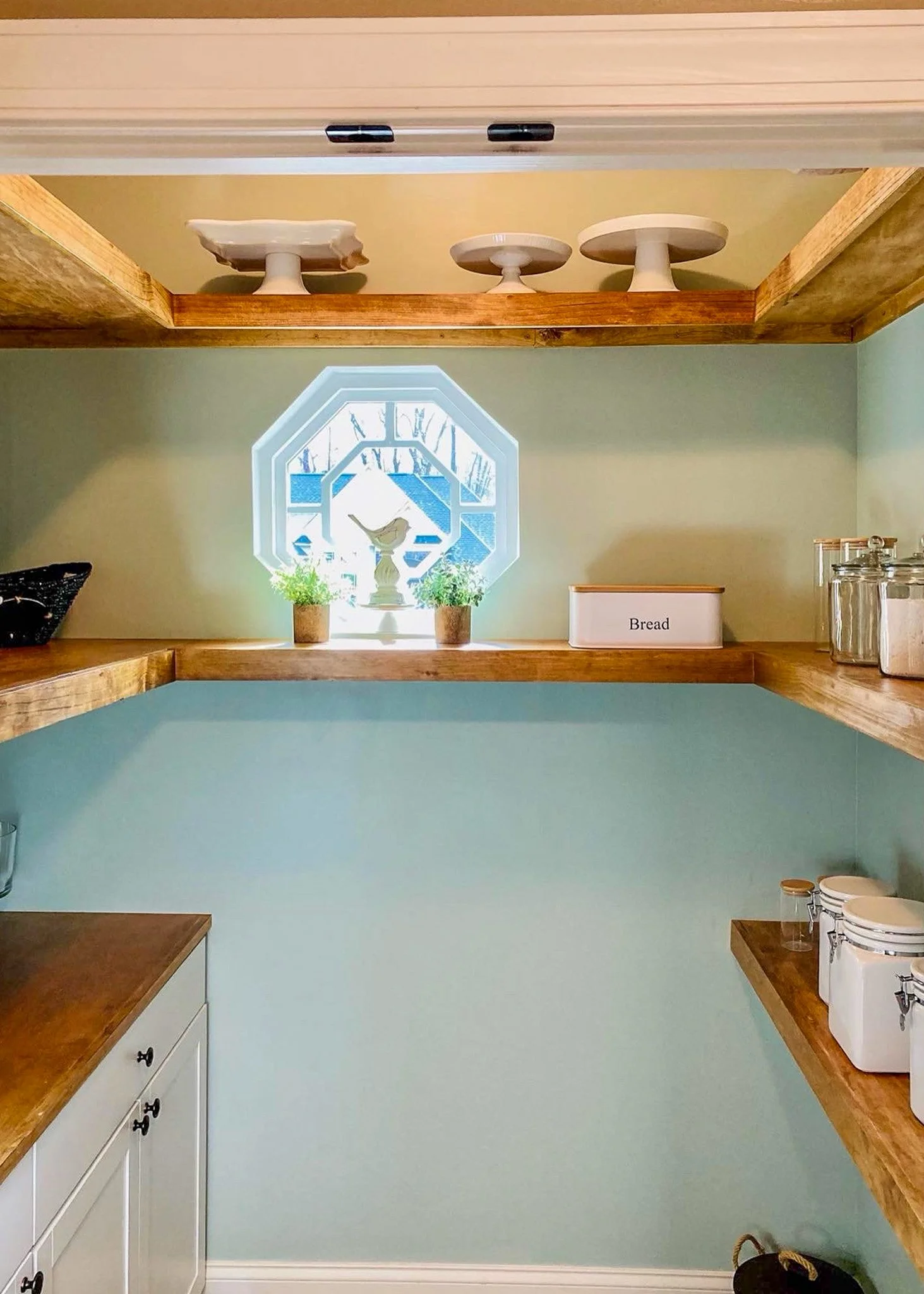 Empty kitchen pantry with wooden shelves, a window with a decorative frame, and labeled containers for bread, jars, and other pantry items.