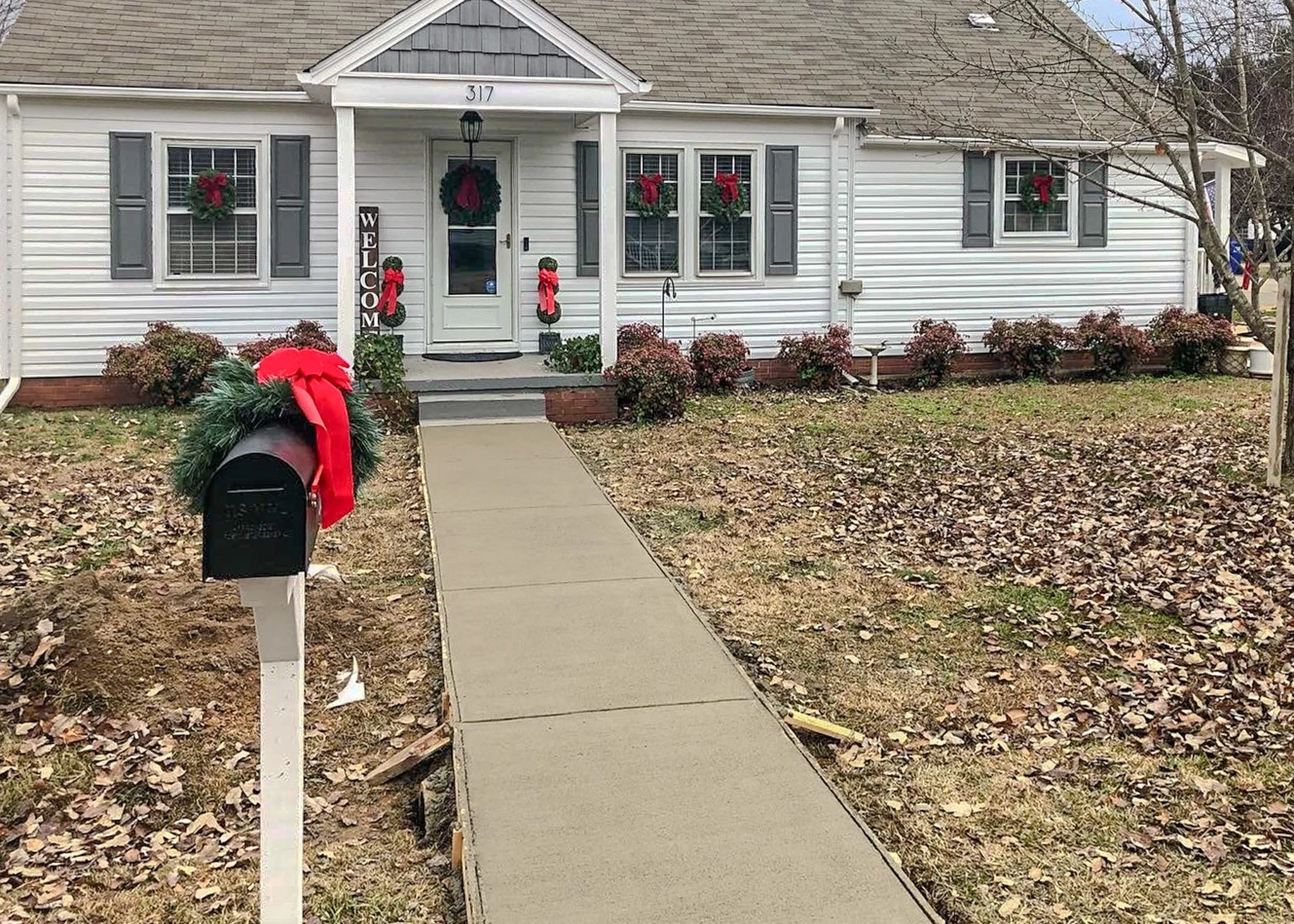 White house decorated for Christmas with red and green wreaths, bows, and a 'WELCOME' sign. A sidewalk leads to the front door, which has a wreath, and a mailbox with a large red bow nearby.