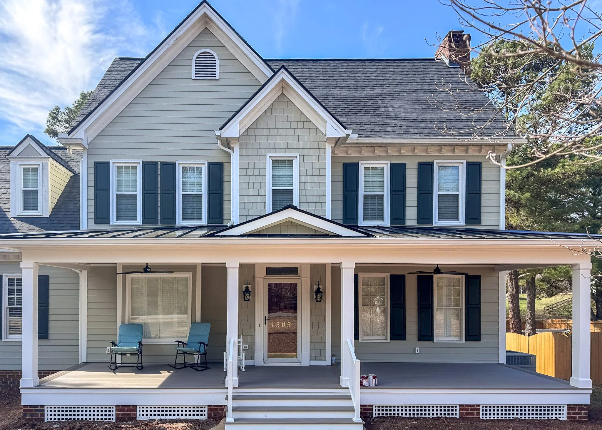 New two-story house with a covered front porch, white pillars, black shutters, and a dark shingle roof. Chairs are on the porch, and a pathway with stairs leads up to the entrance. A wooden fence is visible on the right side, and there are trees and 