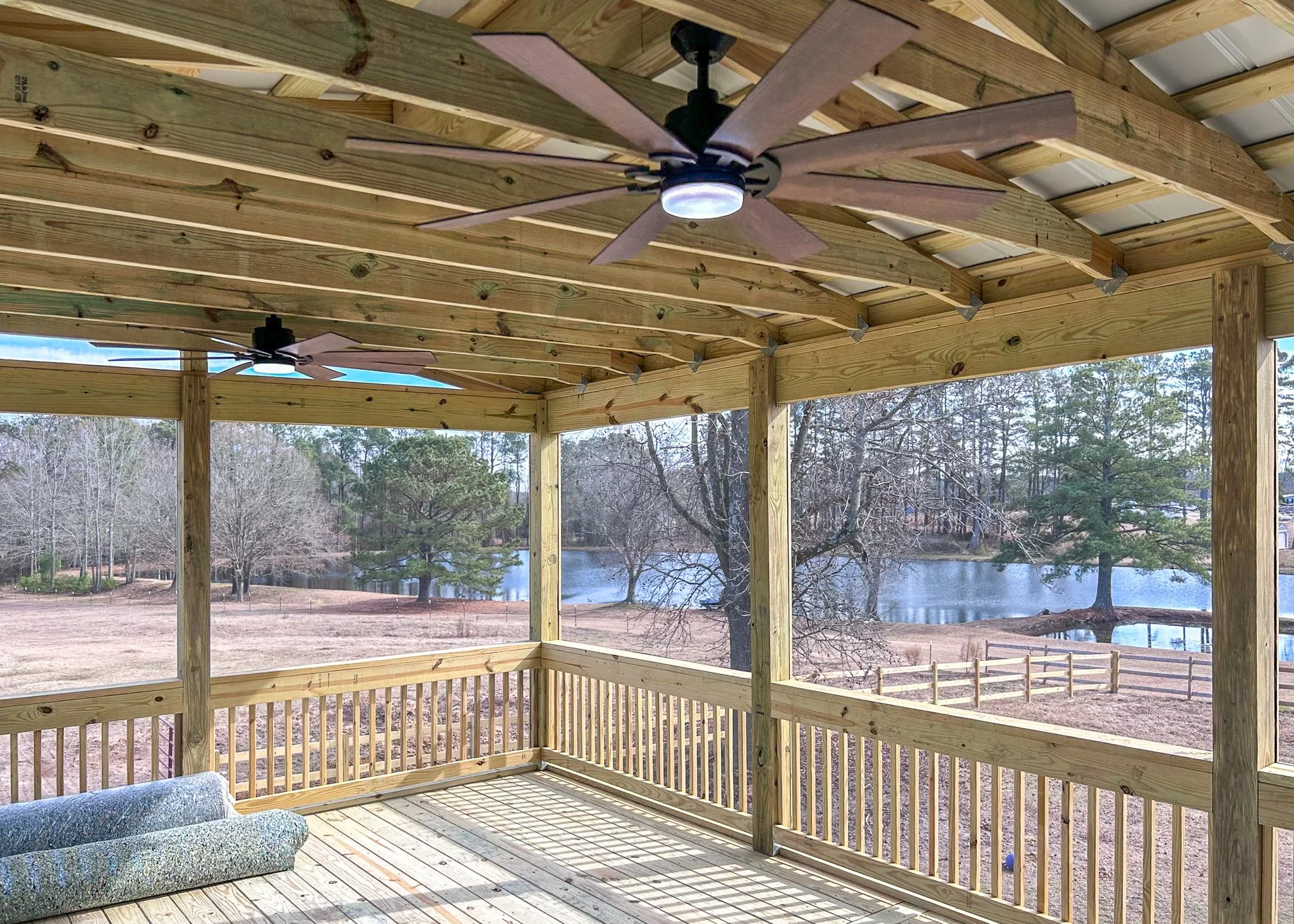 A screened-in wooden porch with ceiling fans, overlooking a lake with trees and a fenced area.