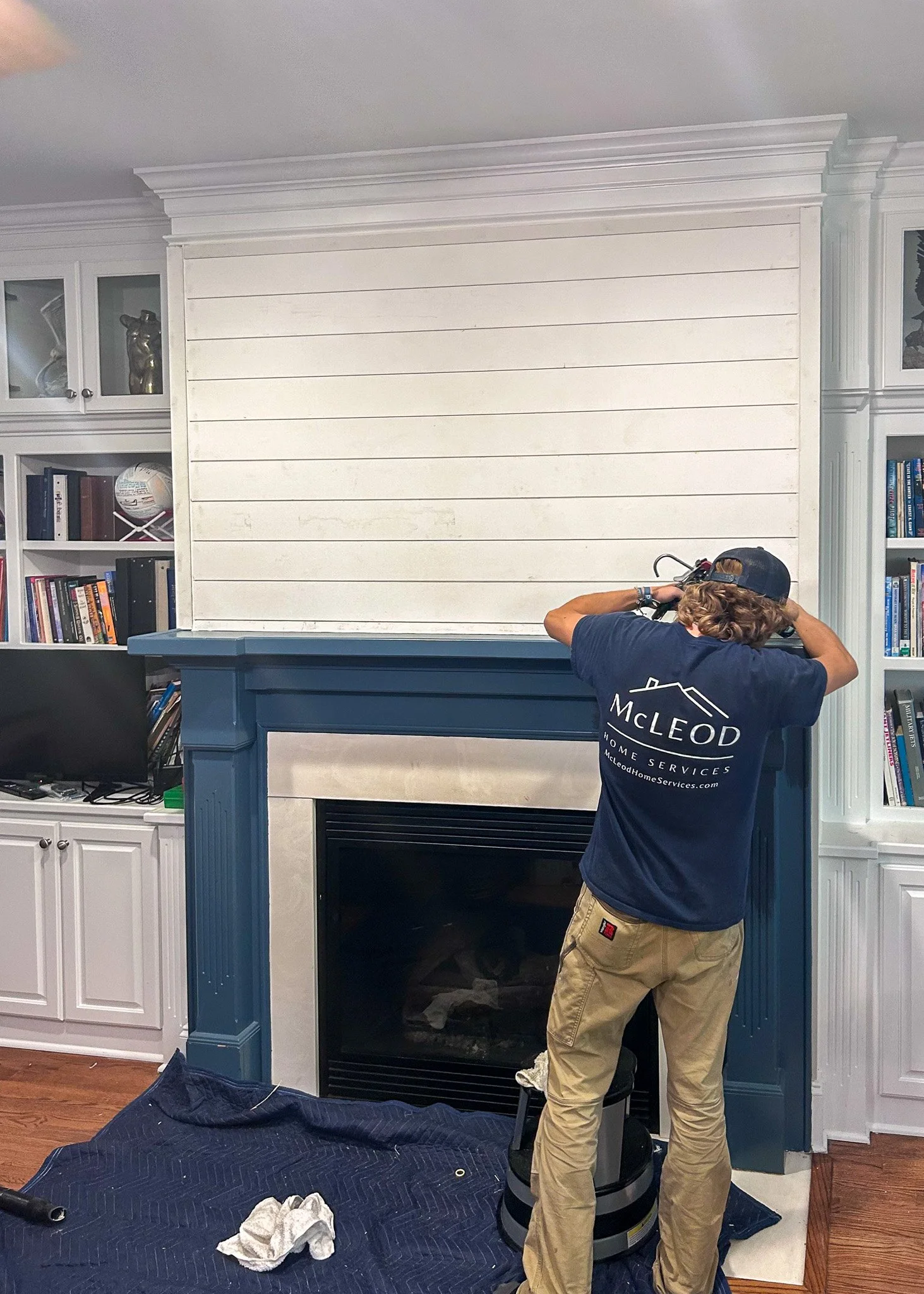 A person working on home improvement in a living room with a fireplace and built-in bookshelves, using tools and wearing a shirt with the logo 'McLEOD.'