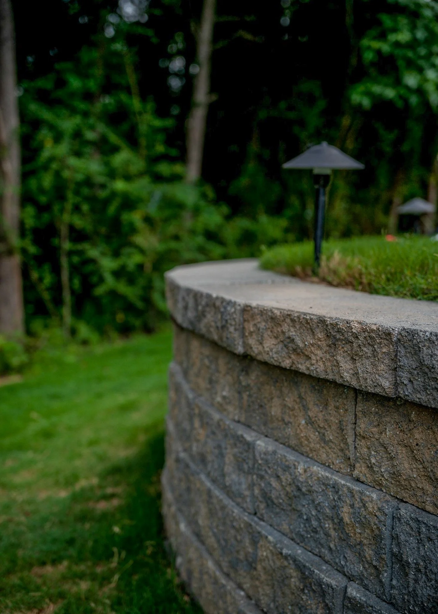 Close-up of a stone wall with a curved edge, topped with a grassy surface and small outdoor lamps, in a wooded outdoor setting.