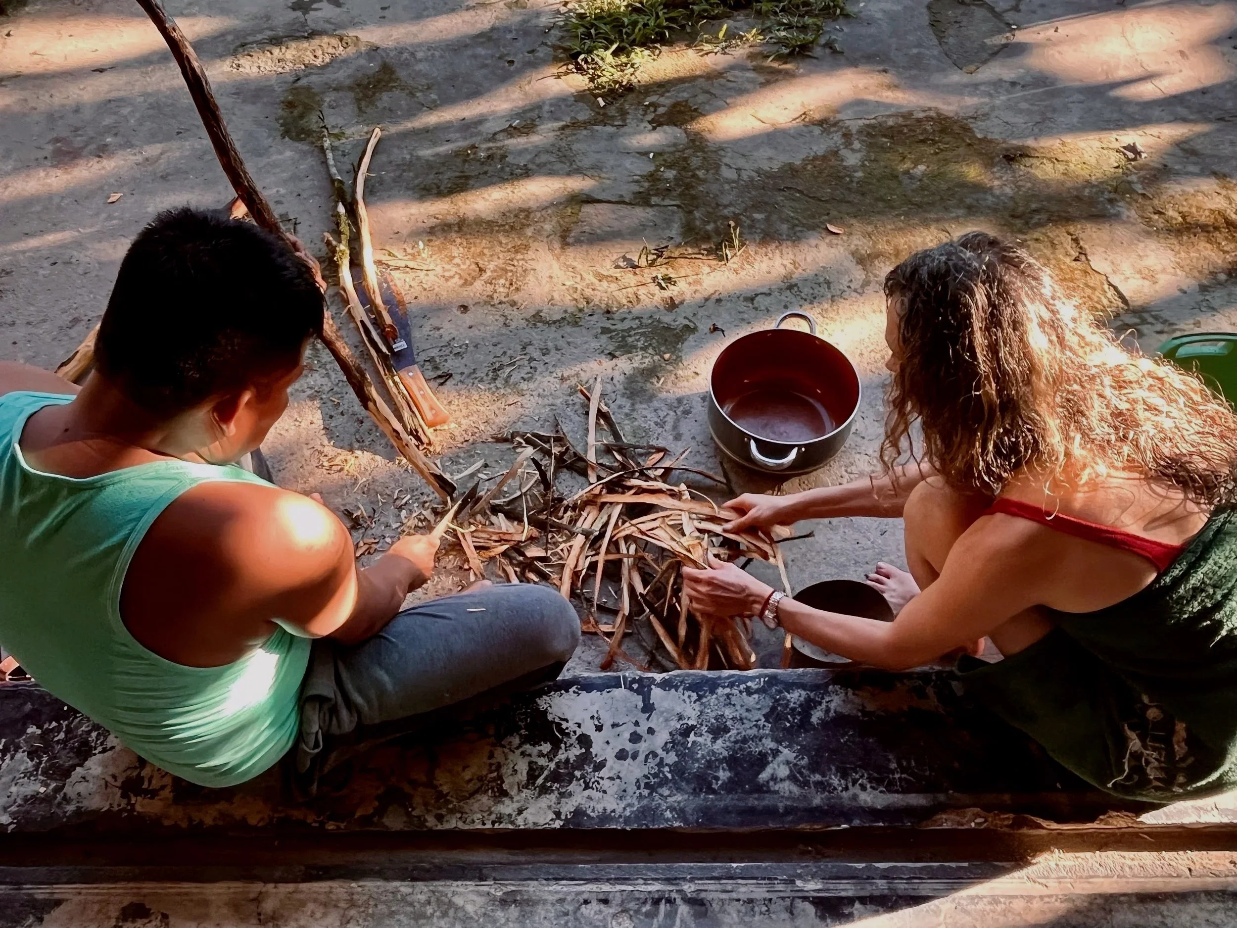 A man and a woman sitting on the ground, stripping bark from branches and placing it into a pot nearby, in an outdoor setting with rocks and foliage.