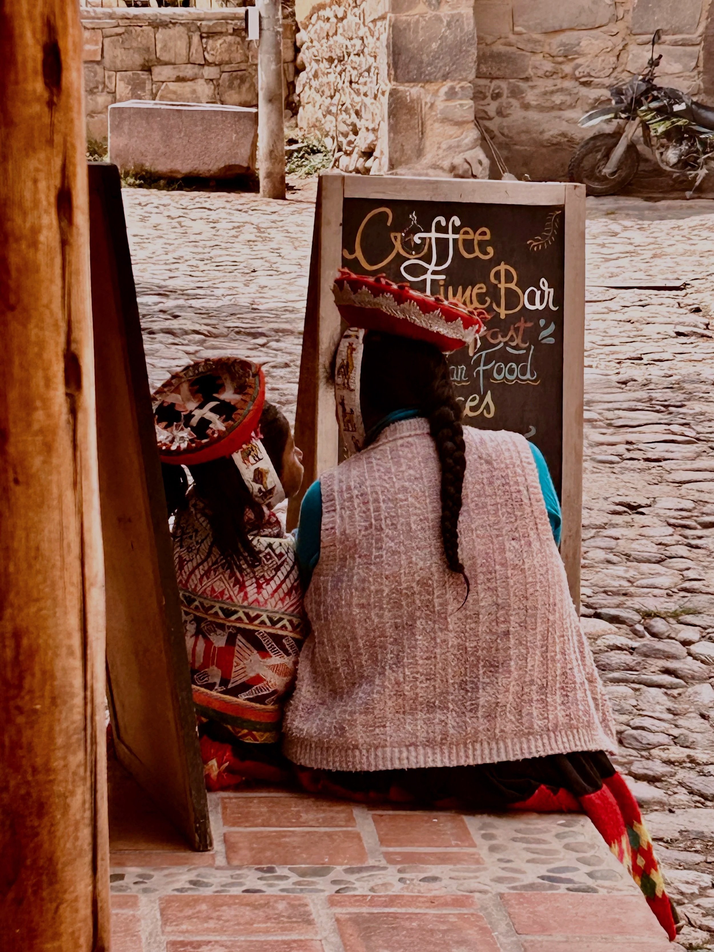 A scene of a mother and child in traditional Peruvian dress and with braided hair, sitting side by side outside a cafe next to a blackboard featuring the word 'coffee'
