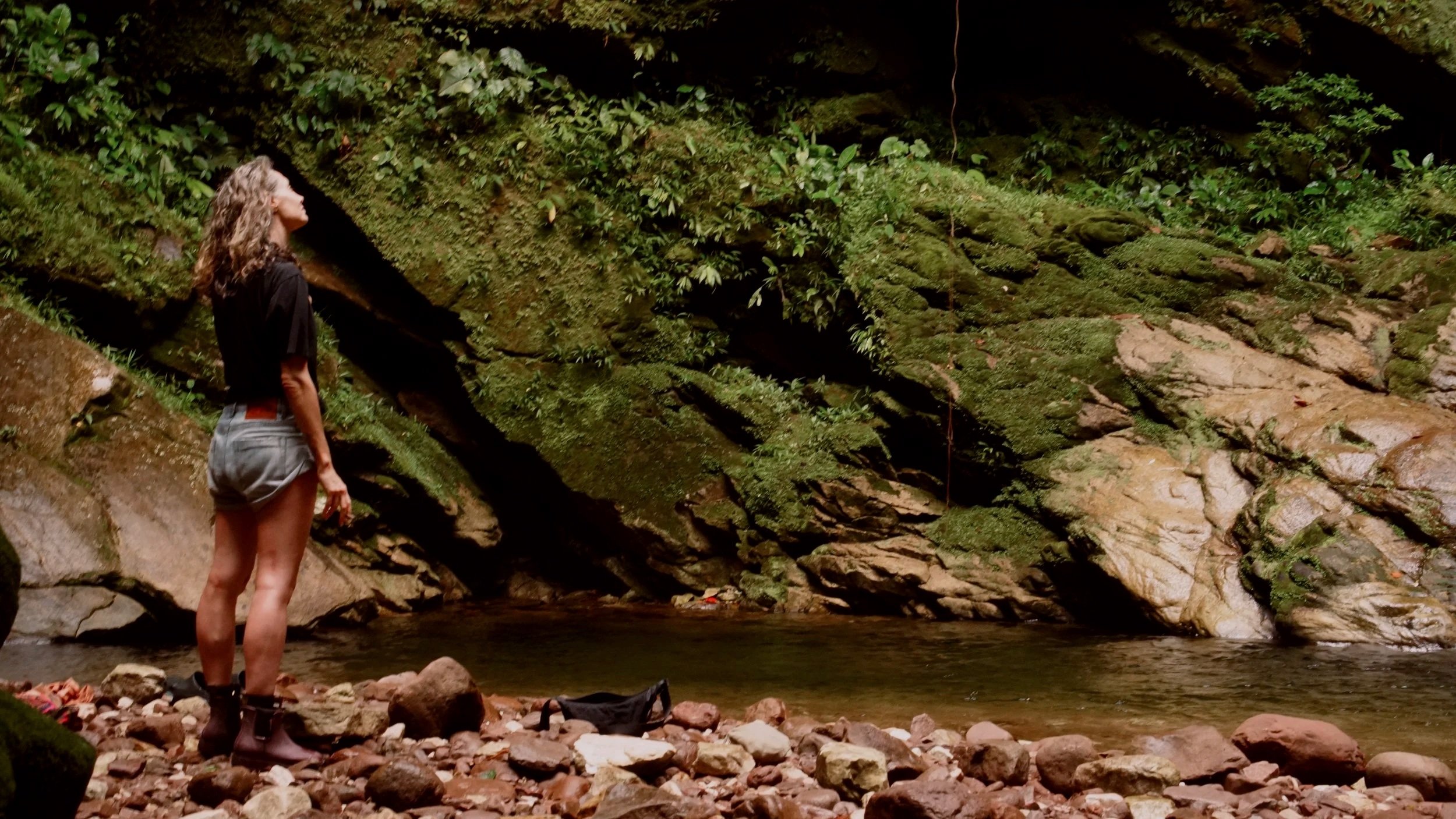A woman with curly hair wearing a black t-shirt, shorts, and boots standing near a rocky riverbank with moss-covered rocks and a small waterfall in the background.