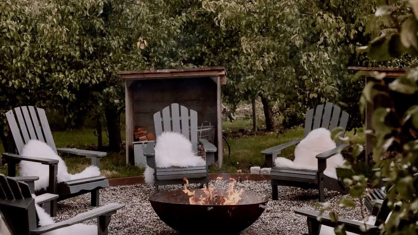 A backyard sitting area with four black wooden Adirondack chairs with white fluffy cushions around a fire pit. There is a small wooden shed behind the chairs. The area is surrounded by trees and greenery.