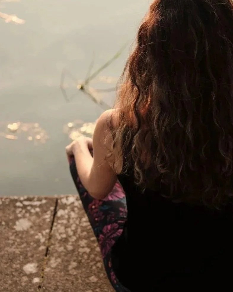 A woman with curly brown hair and wearing a black top and floral leggings, sitting by a body of water, fishing with a fishing rod.