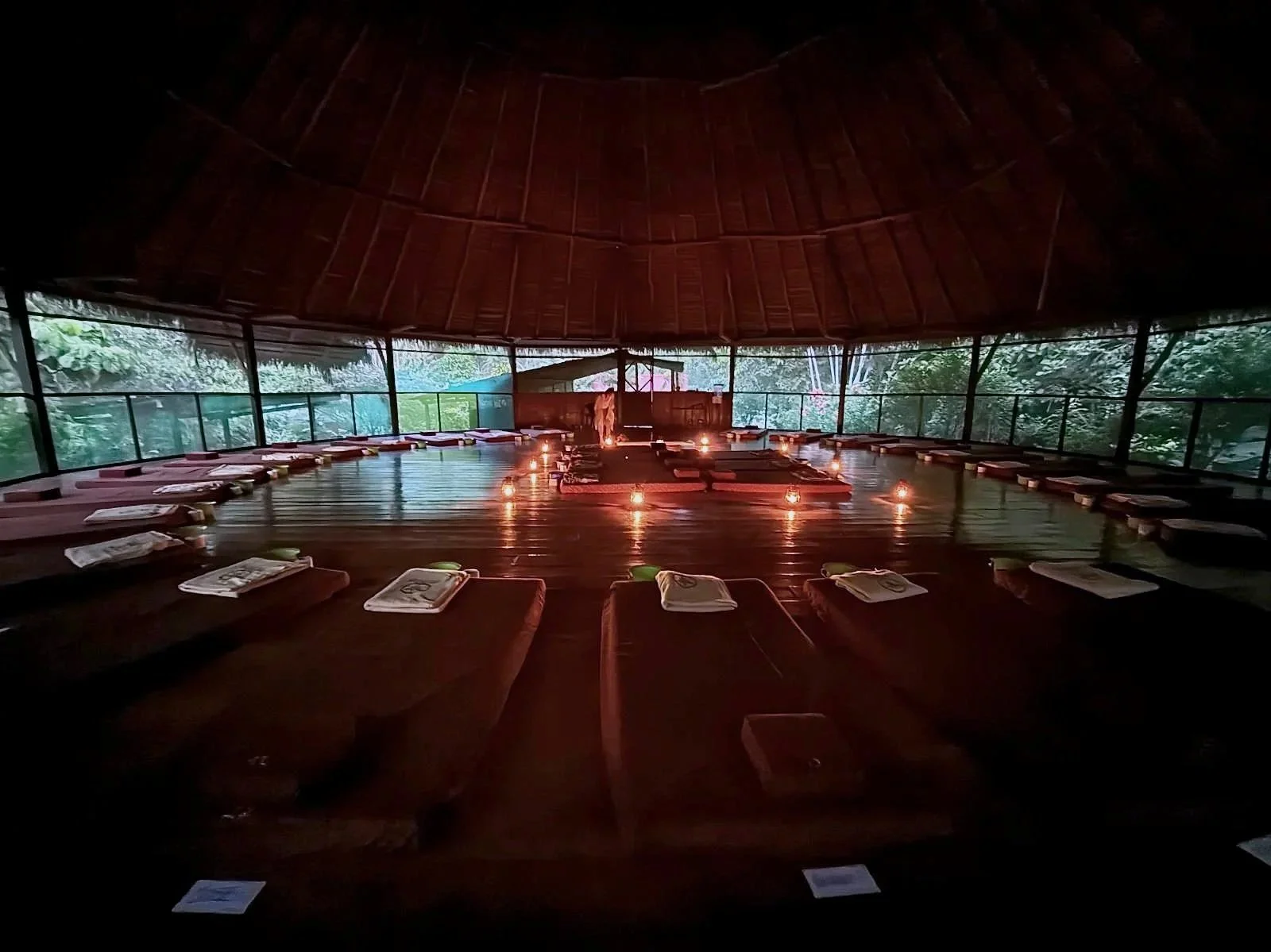 A spacious, open-air, circular ceremonial space or maloca with a thatched roof, and with mattresses and cushions arranged in a circle, surrounded by lush greenery, with candlelight creating a calming atmosphere.