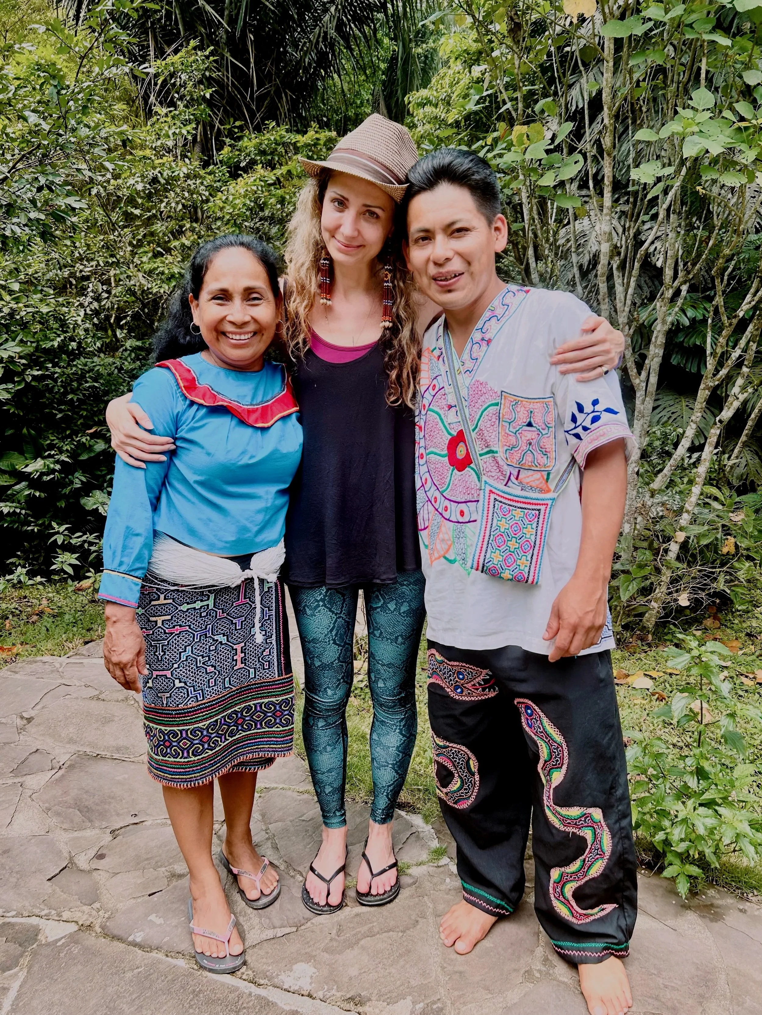 Three people standing outdoors in front of green foliage, smiling and posing for a photo. The group includes two women and one man, all dressed in colorful traditional and modern attire, with the woman on the left wearing a blue top and patterned skirt, the woman in the middle wearing a black dress with a hat and leggings, and the man on the right wearing a white embroidered shirt and black pants.