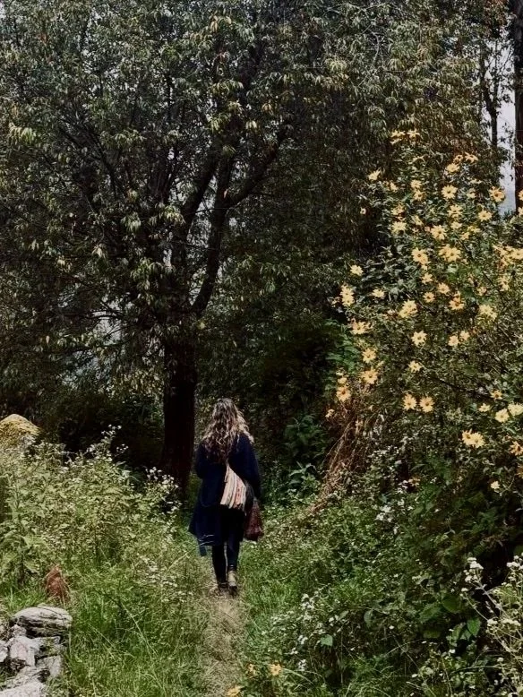 A woman walking along a narrow dirt trail surrounded by lush greenery and flowering bushes, with large trees and dense foliage in the background.