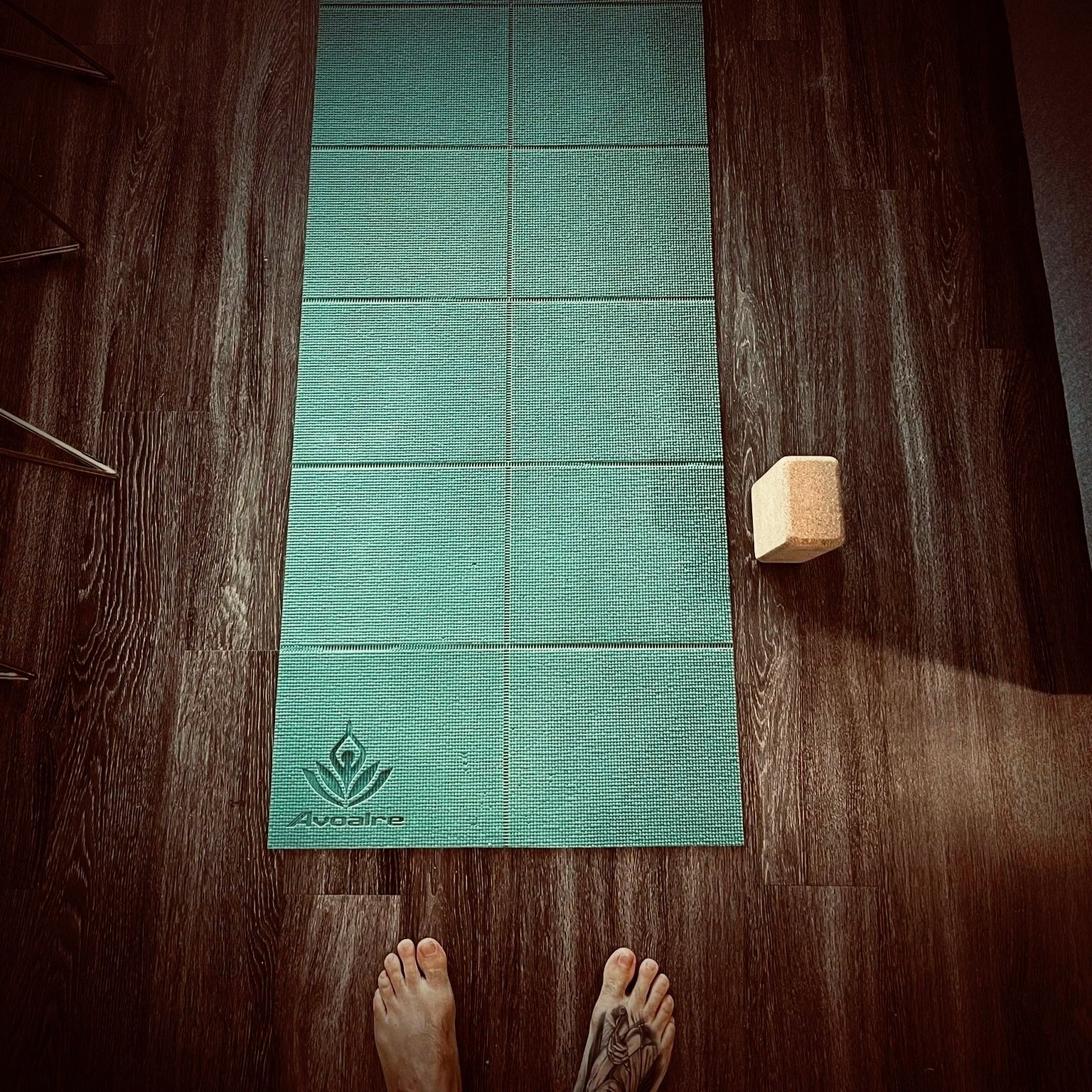 A person standing on a wooden floor, looking at a yoga mat, with a cork block, and part of a chair on the left side.