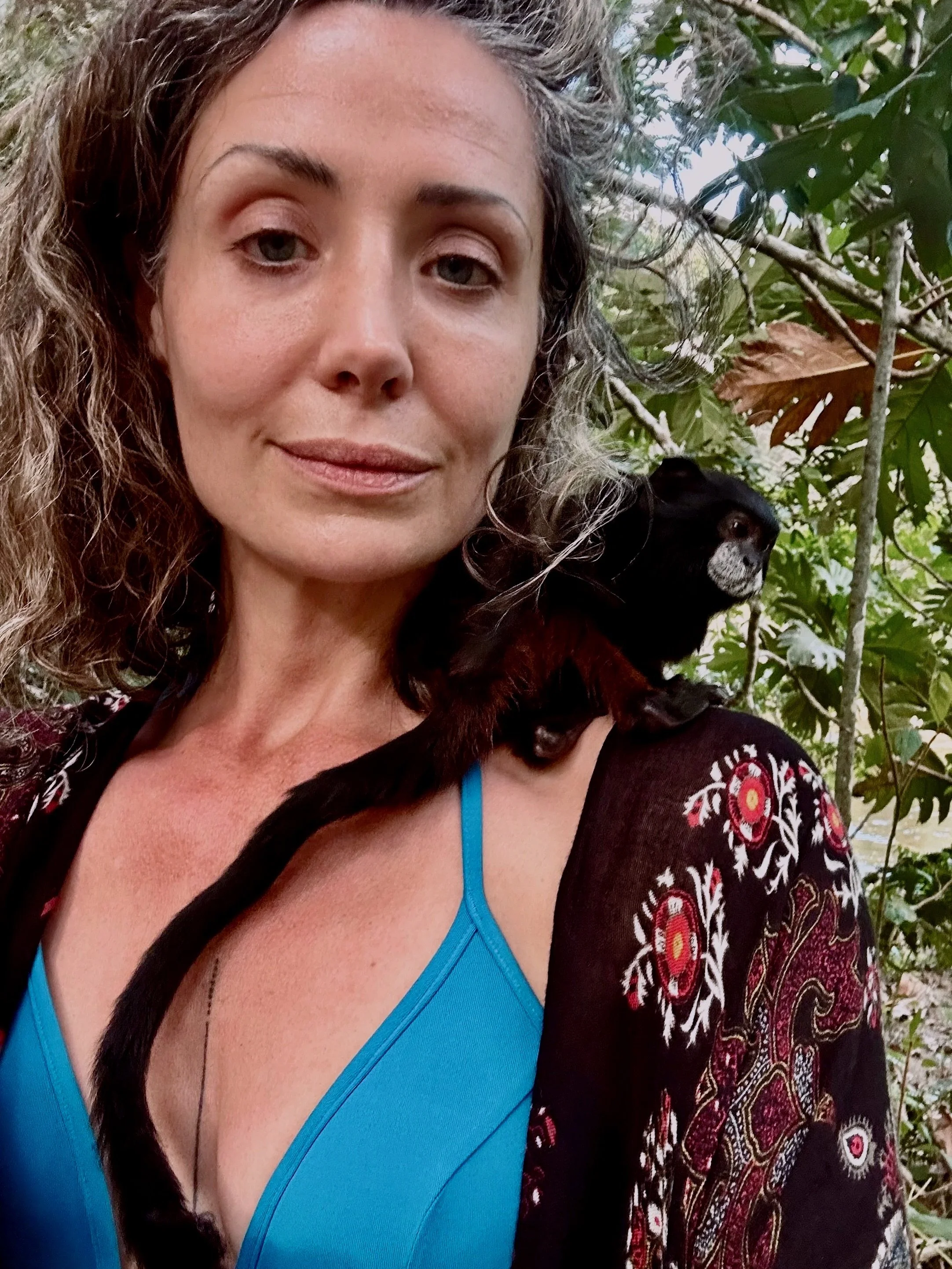 A woman with curly hair taking a selfie outdoors among green foliage, with a small monkey sitting on her shoulder.
