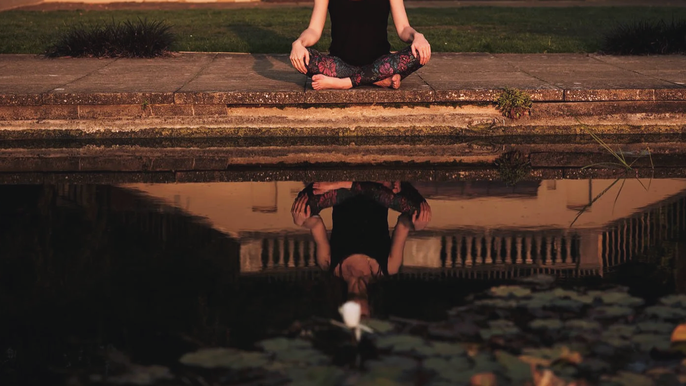 Person sitting cross-legged on the sidewalk practicing meditation at sunset, with their reflection visible in a pond below.