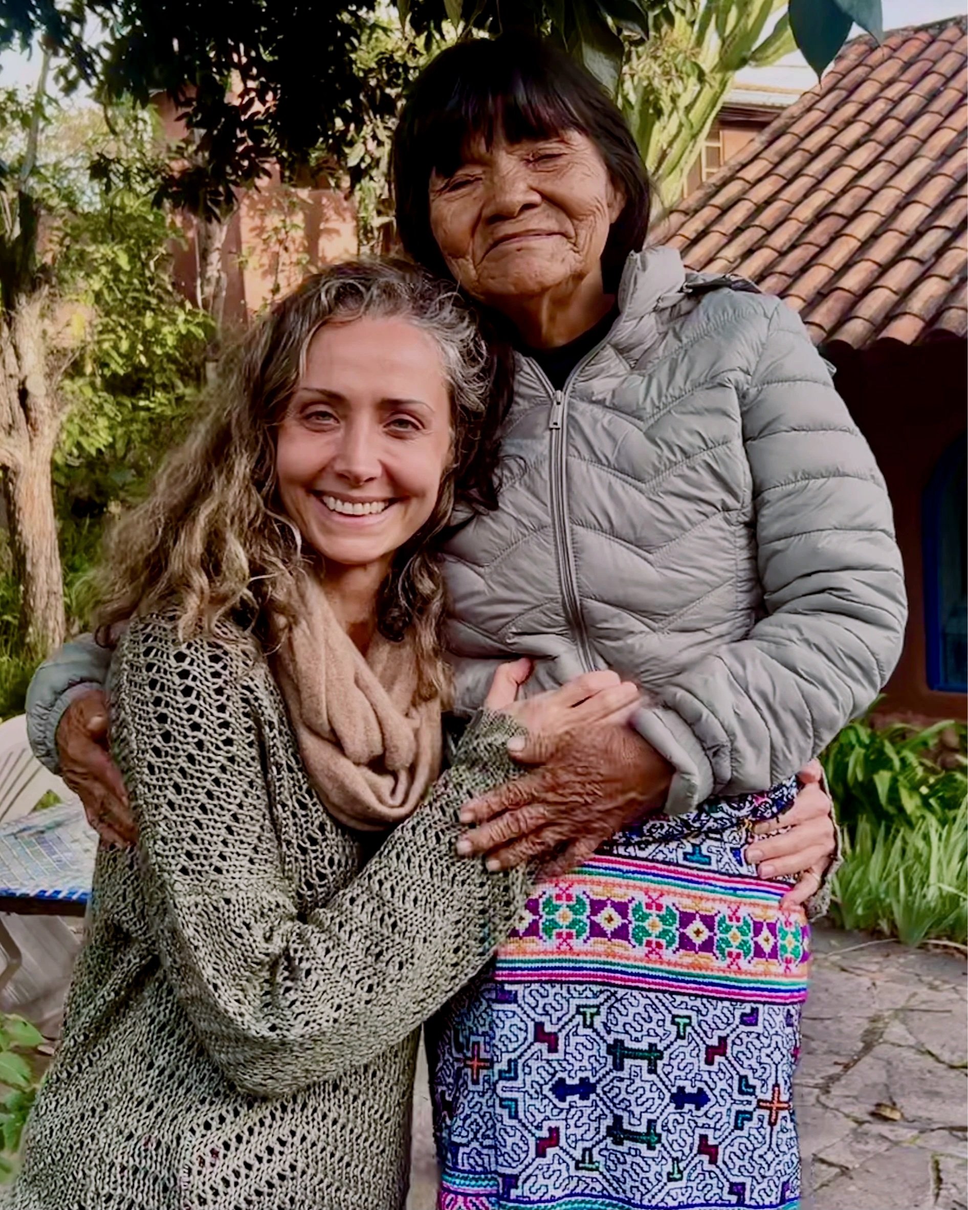 Two women hugging outdoors, one appears to be an elderly indigenous woman with dark hair and others appear middle aged with curly hair, wearing various clothing styles, standing in front of greenery and a house with a tiled roof.