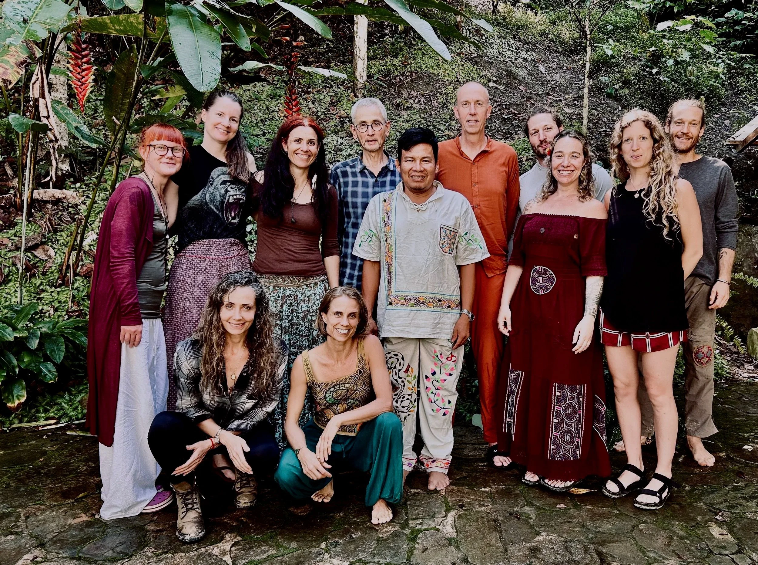 A group of diverse retreat participants standing and sitting together outdoors in a lush green forest, smiling at the camera.