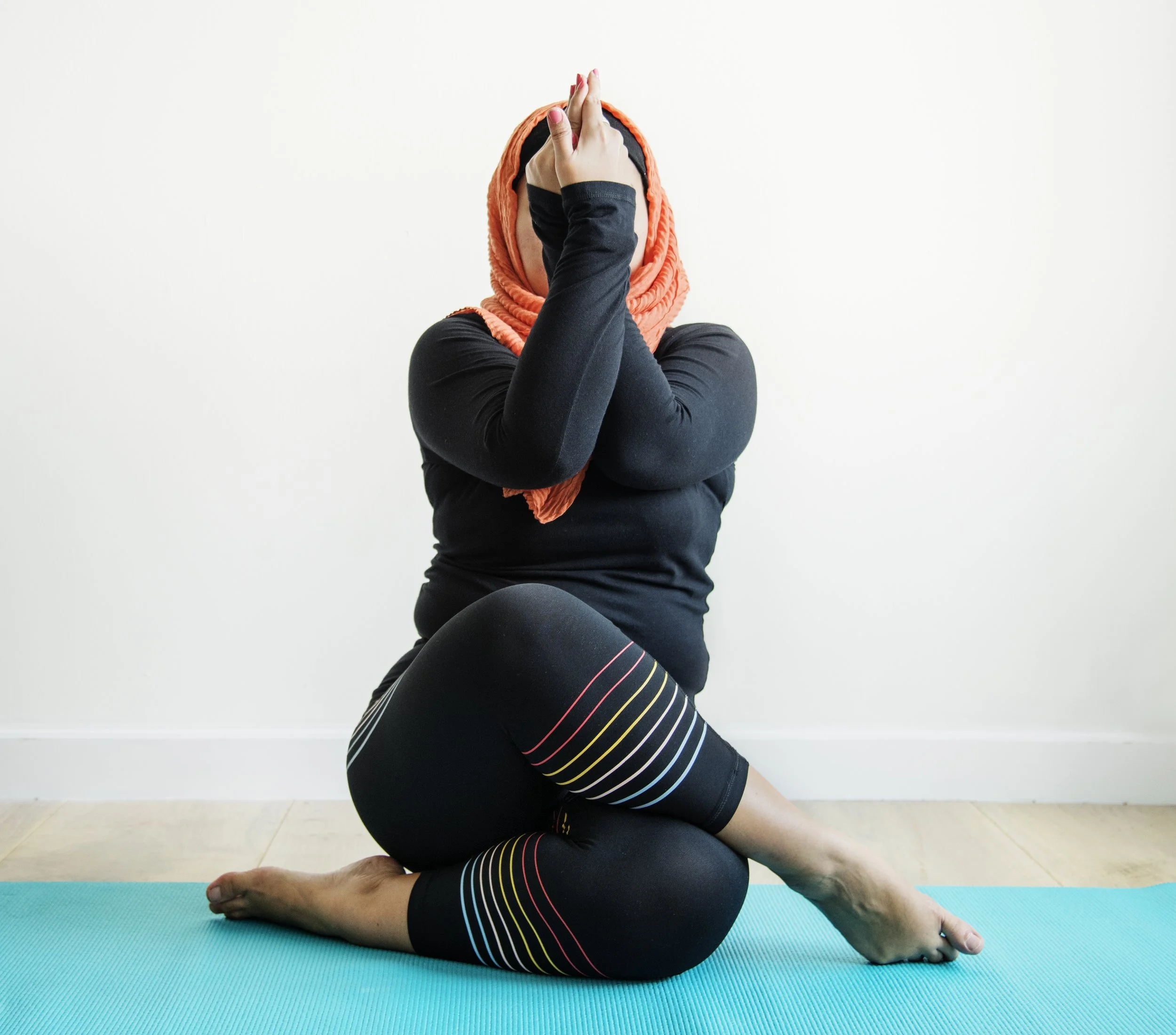 Person in modest clothing and a headscarf practicing yoga on a blue mat indoors, sitting with one leg crossed and hands pressed together in a prayer position near the face.