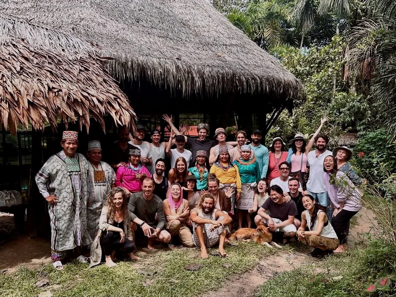 A diverse group of people smiling and posing for a photo outside a traditional palm thatched ceremonial hut in a lush, green, Amazonian jungle setting, with some dressed in traditional Peruvian attire.