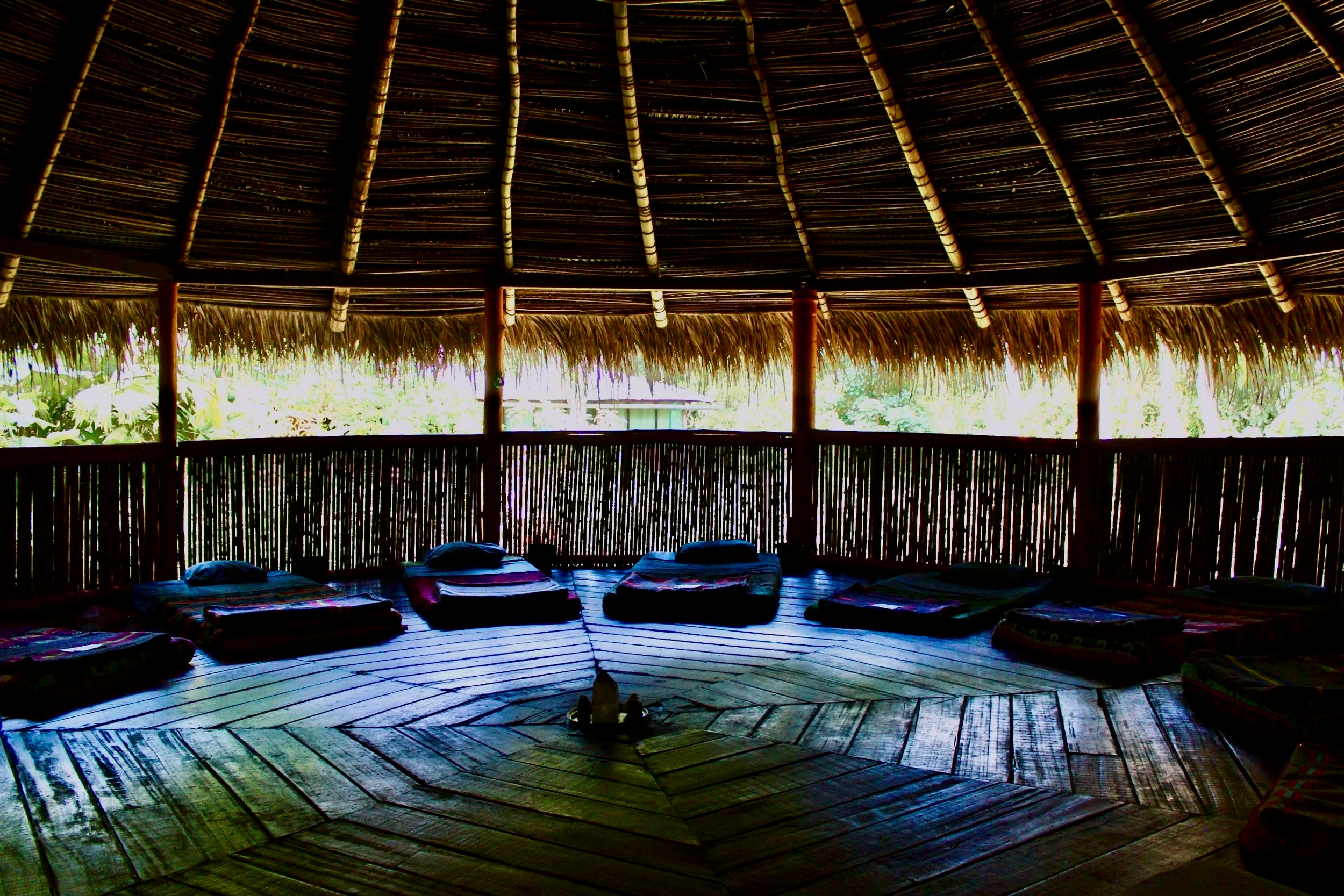 Interior of a circular ceremonial hut with thatched roof, wooden floor and platforms, with mattresses and pillows, overlooking lush greenery outside.