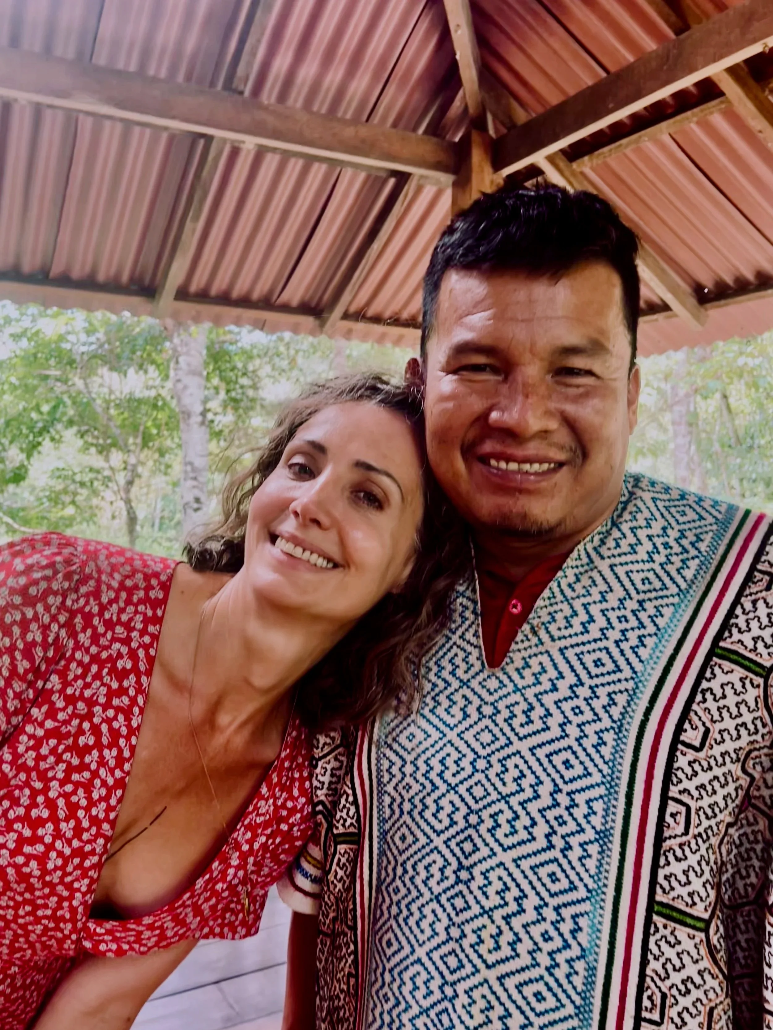 Close-up of two smiling people, one woman and one man, the man in indigenous Peruvian clothing indoors with a wooden ceiling and trees in the background.