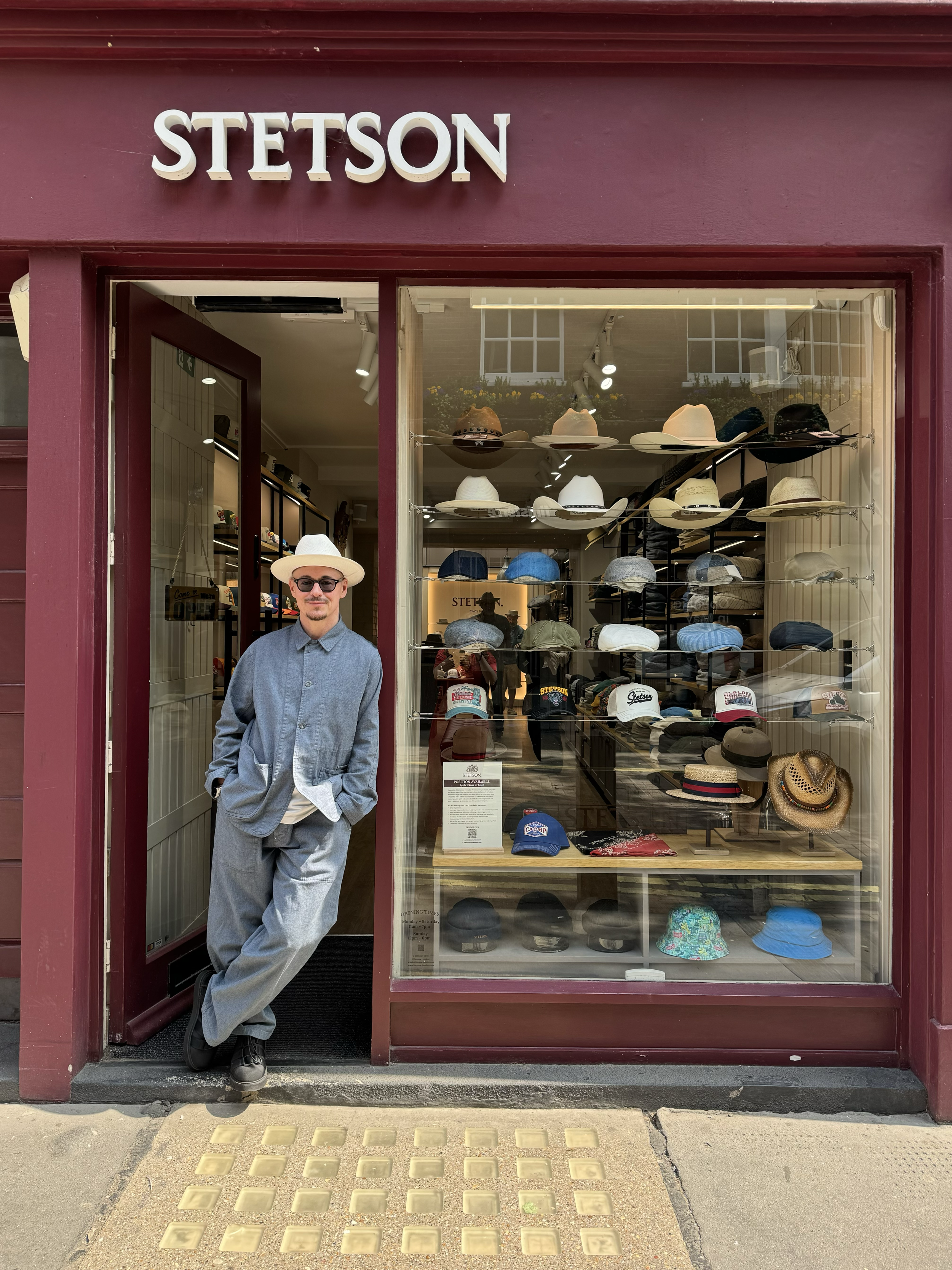 Man in hat and sunglasses standing outside a Stetson hat store with various hats displayed in the window.