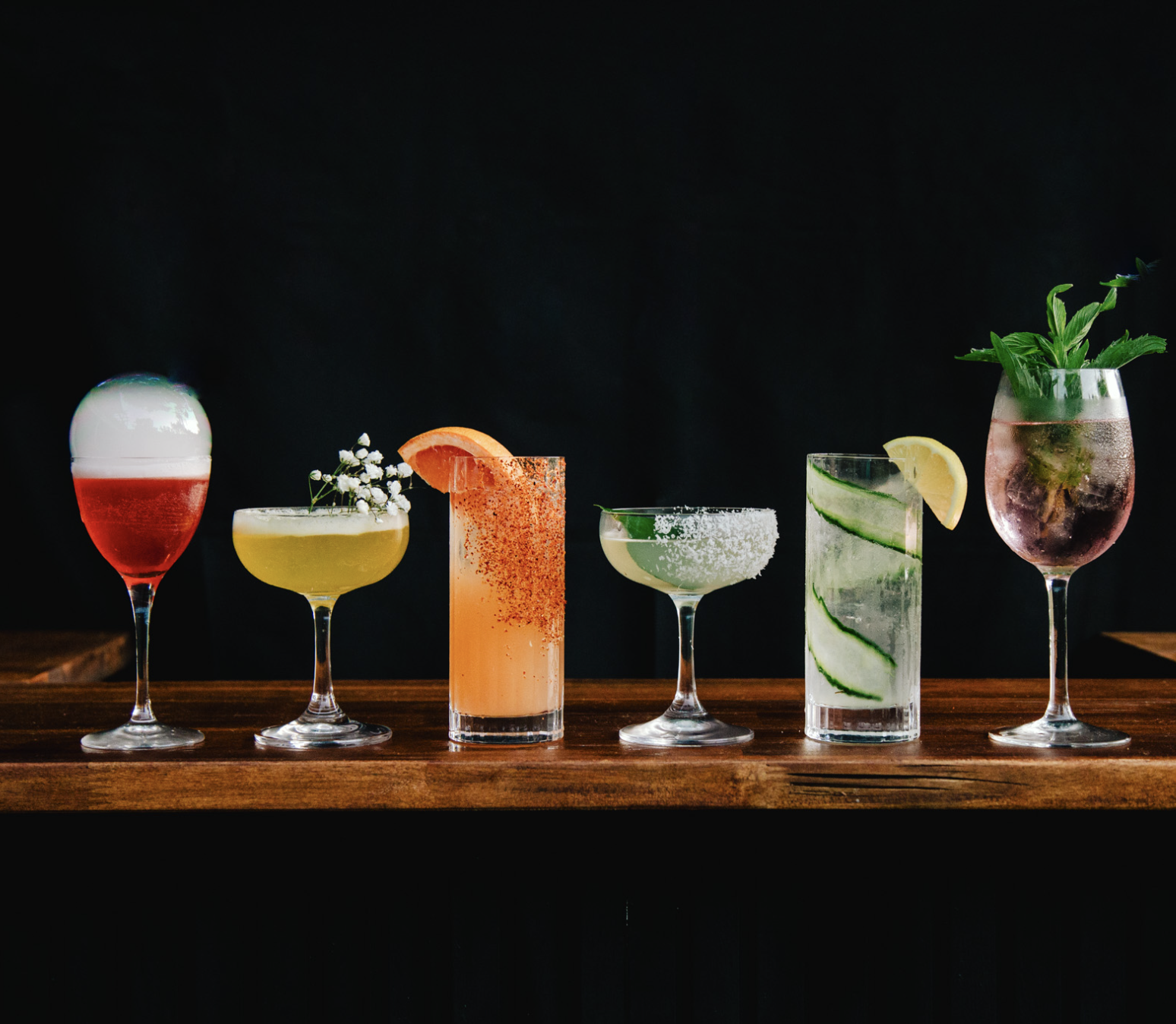 Six colorful cocktails on a wooden bar counter against a black background.