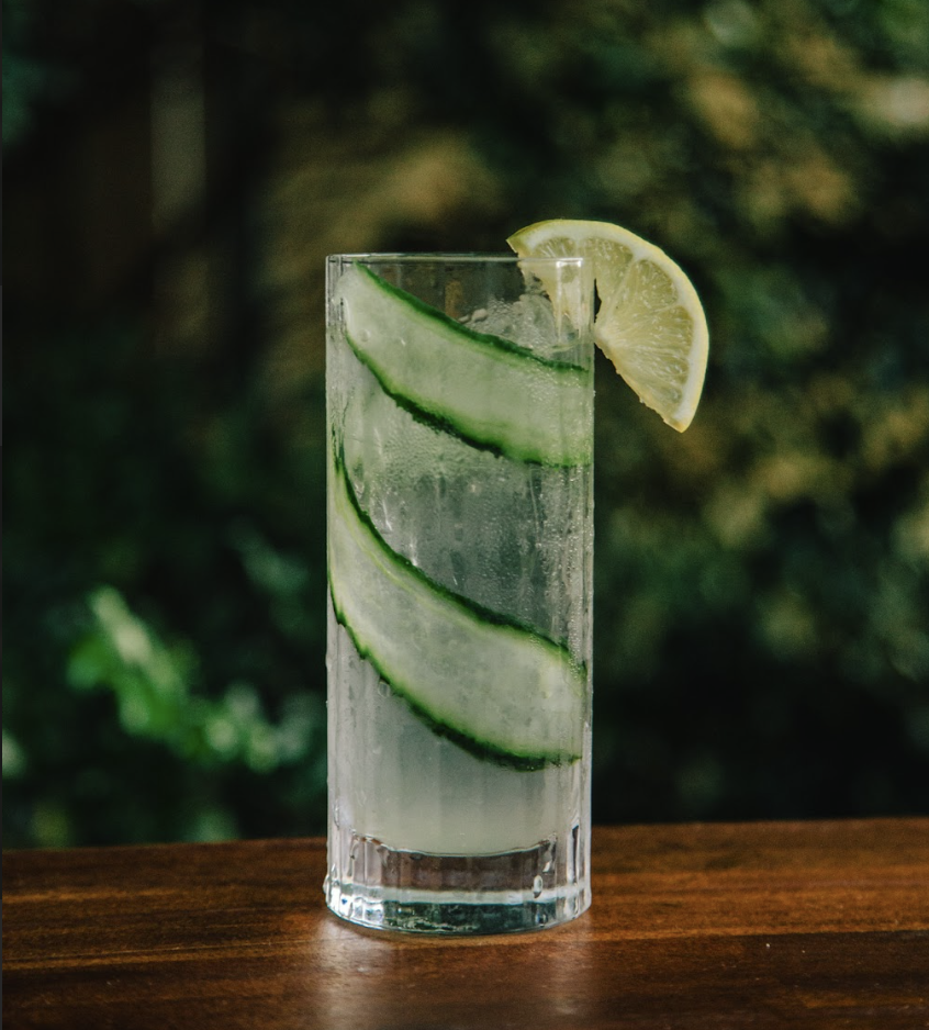 A tall glass of clear fizzy water with cucumber slices and a lemon wedge garnished on the rim, sitting on a wooden surface.