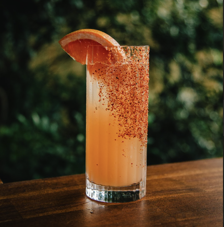 A tall glass filled with a pink-orange beverage, garnished with a slice of grapefruit on the rim and sprinkled with seasoning or chili powder.