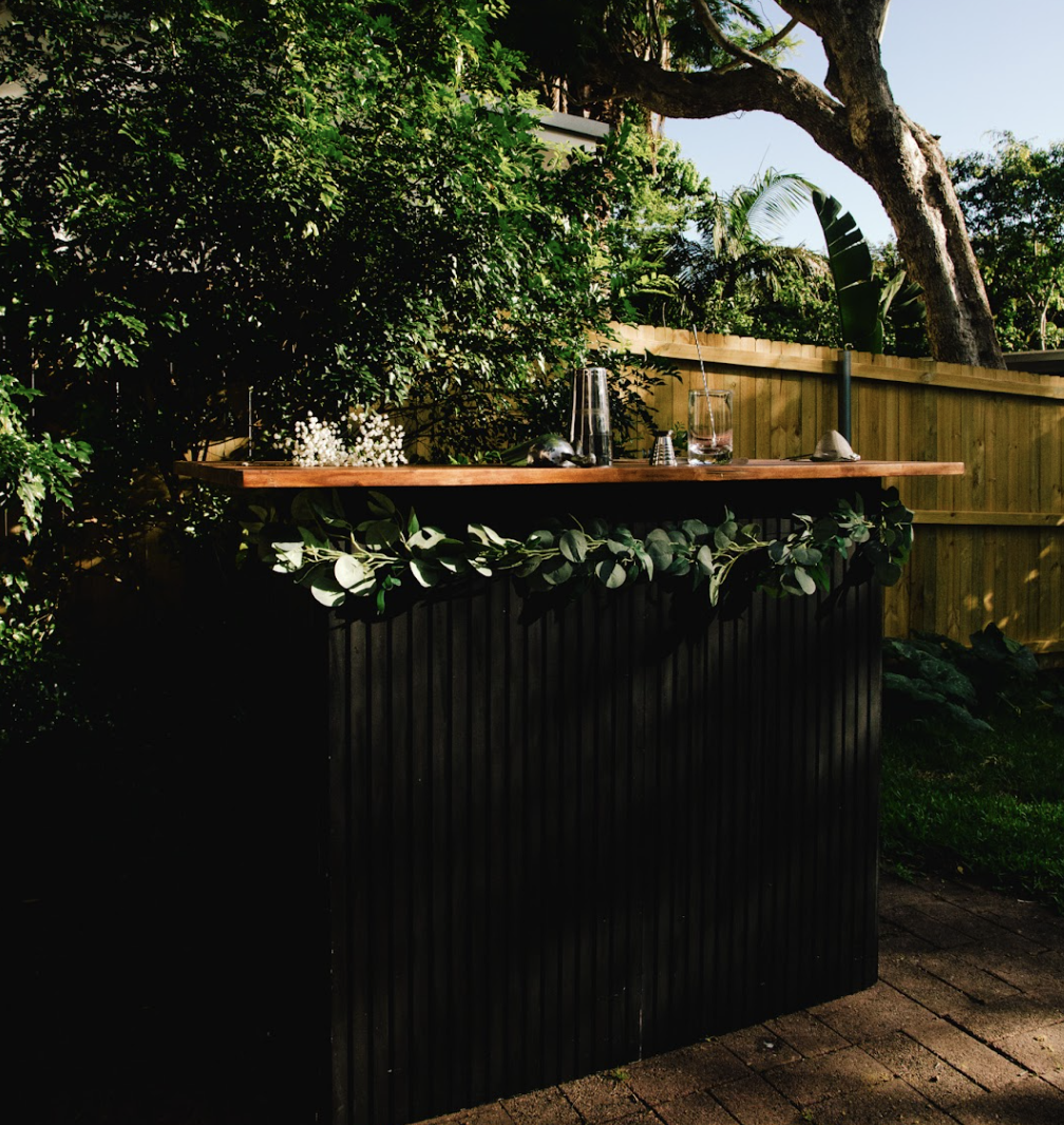 Outdoor bar with a wooden top and black base, decorated with green leaves, set in a lush backyard with trees, bushes, and a wooden fence.