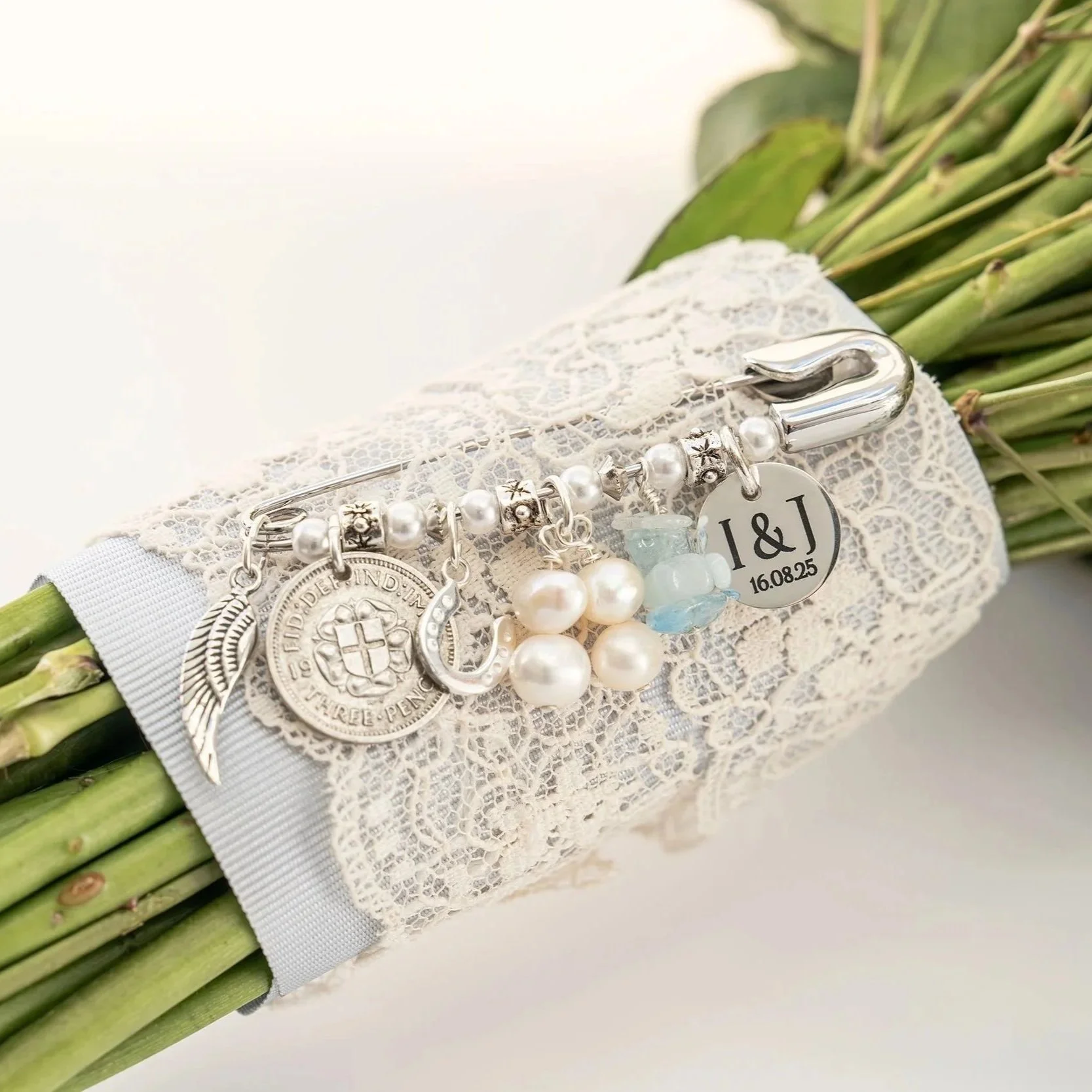 A keep sake Bridal Pin Charm featuring something old, new, borrowed and blue is attached to a bouquet of flowers which has the stems wrapped in lace and pale blue ribbon. A monogram engraved disc with the couples initials and date of wedding is featu