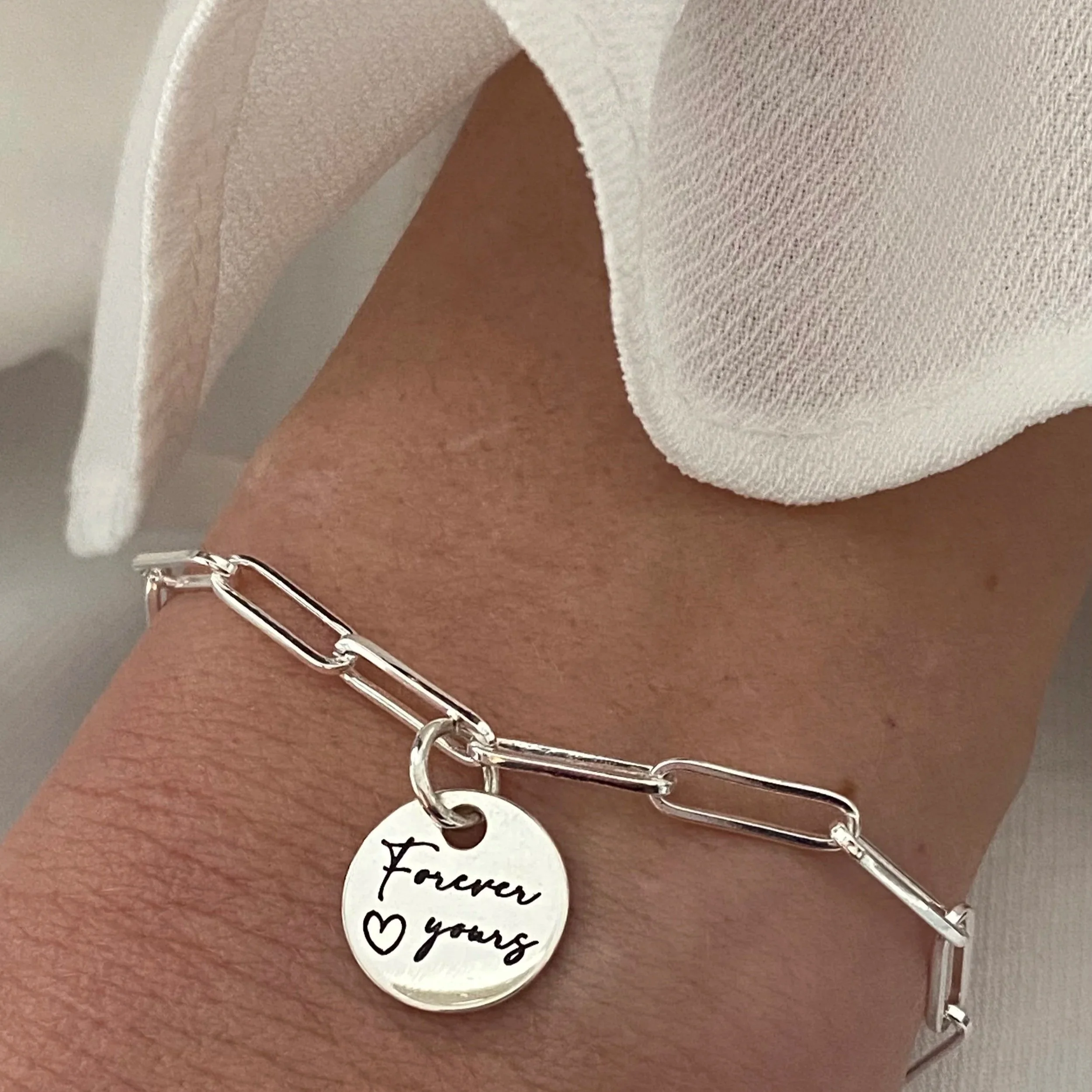 Silver bracelet with circular charm inscribed "Forever yours" and a heart, worn on a wrist.