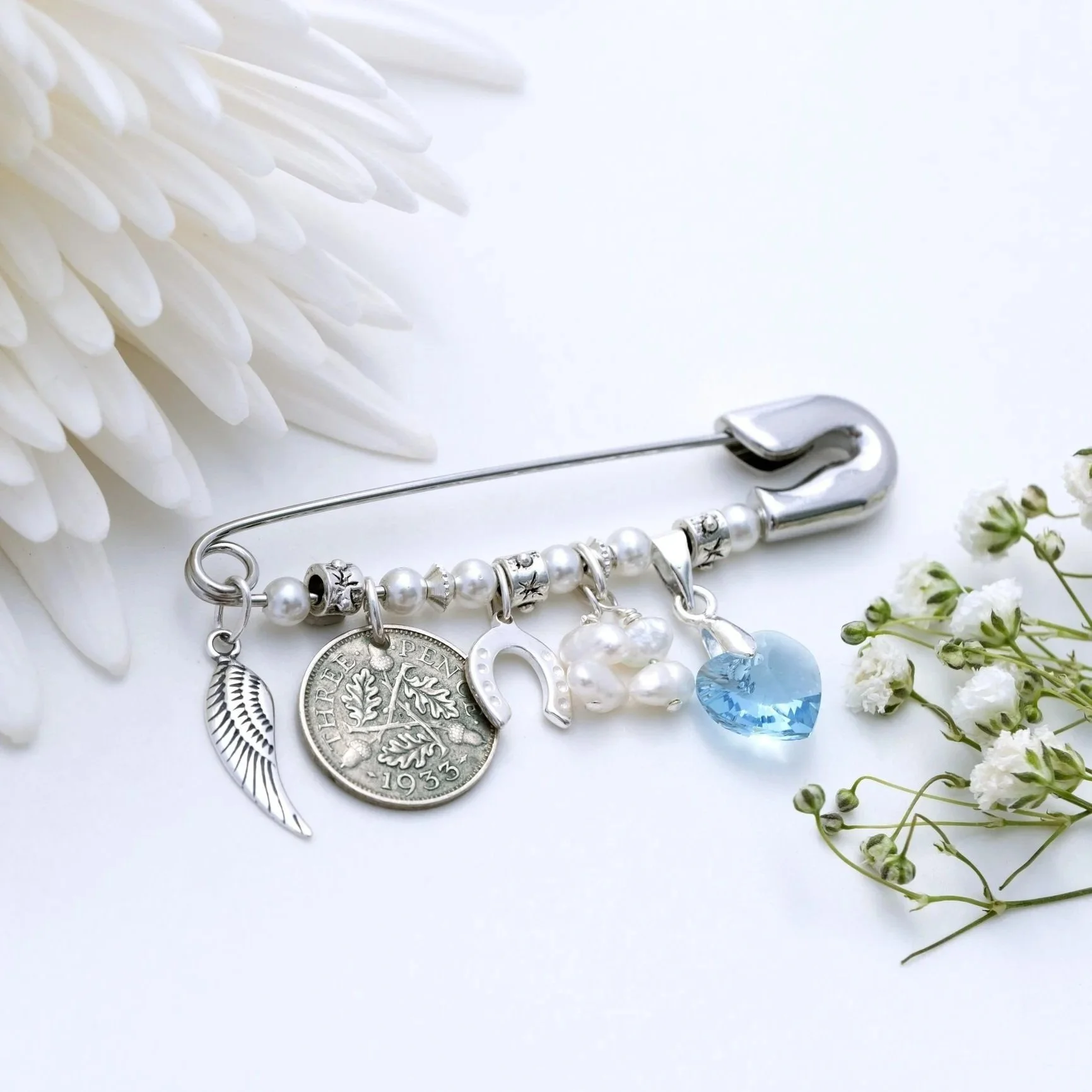 A bridal bouquet pin with charms including an angel wing, threepence coin, a silver horseshoe, freshwater pearls, blue heart-shaped swarovski crystal pendant, placed between white flowers on a light background.