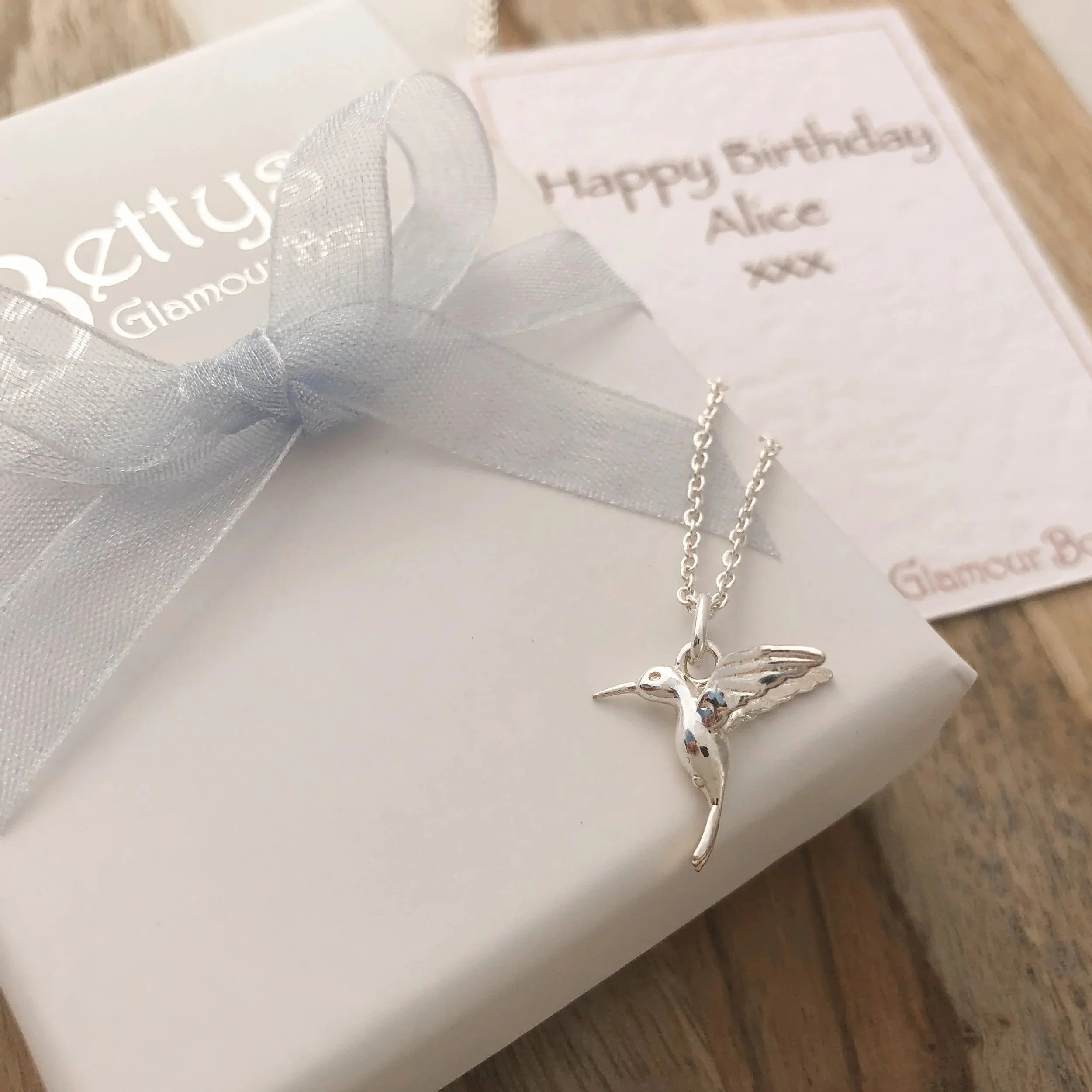 Silver hummingbird necklace on a gift box with a ribbon and a birthday card in the background.