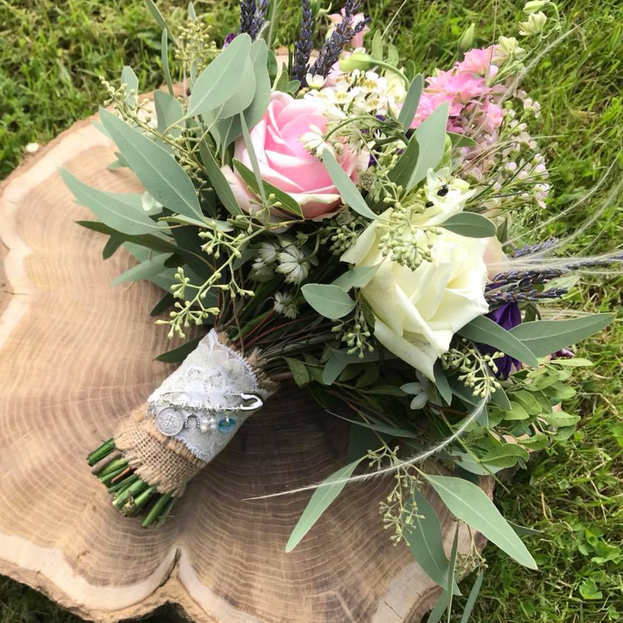 Bouquet of pink and white roses with greenery and lavender, placed on a tree stump with a bridal bouquet good luck charm attached to the wrap.