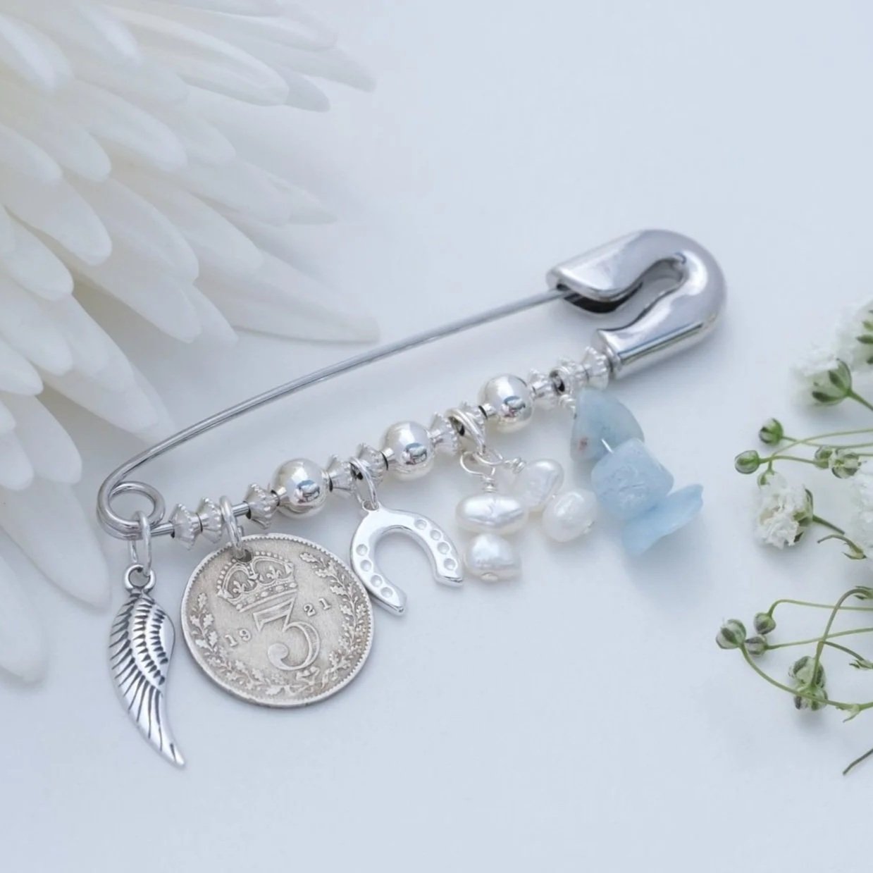 Close-up of a decorative pin adorned with charms including an old coin, wing, horseshoe, and pearls, aquamarine stones, placed between white flowers on a light background.