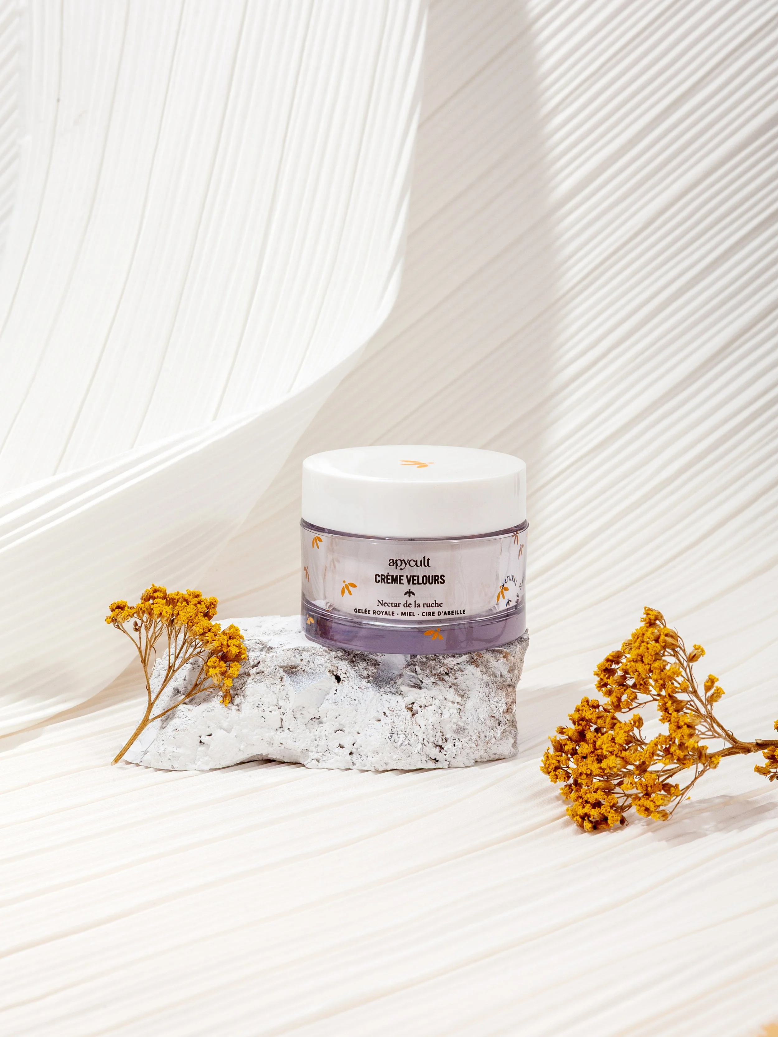 Pot de crème pour les lèvres posé sur une pierre, entouré de fleurs jaunes, avec un fond blanc lumineux.