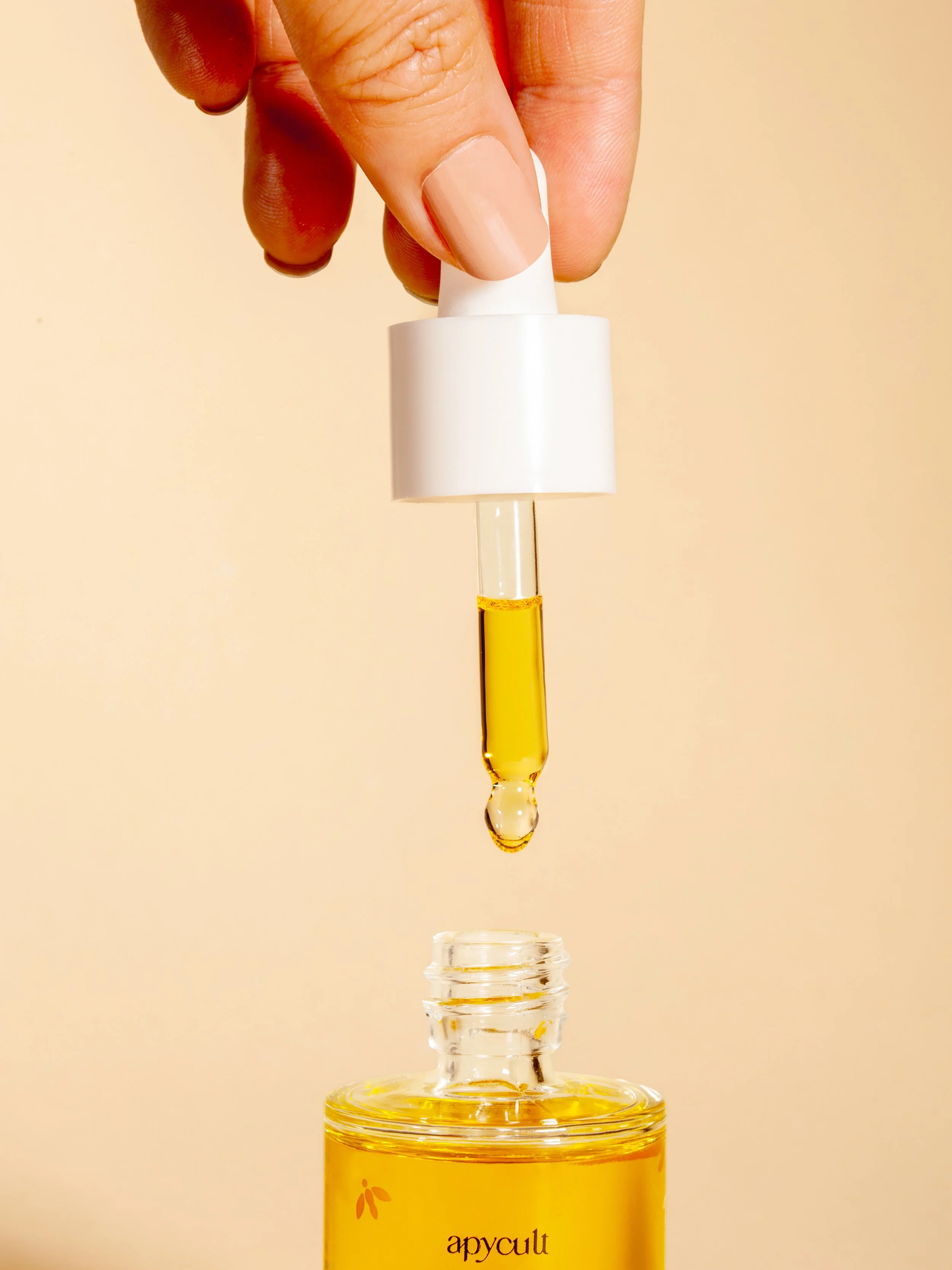 Une main tient un flacon compte-gouttes avec de l'huile jaune, en train de déposer quelques gouttes dans un flacon en verre.