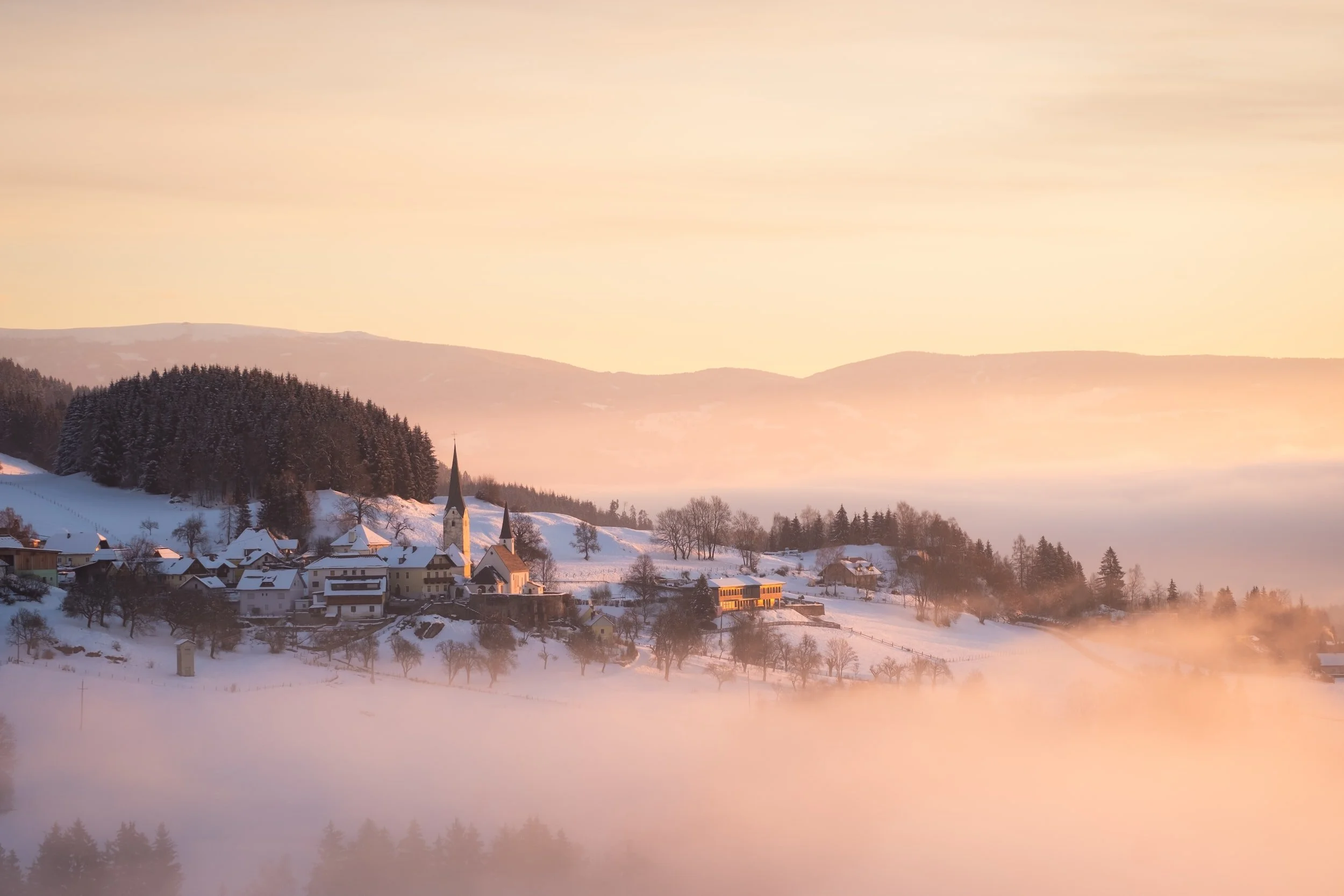 Snow-covered village at sunrise with church and trees, surrounded by mountains and misty sky.