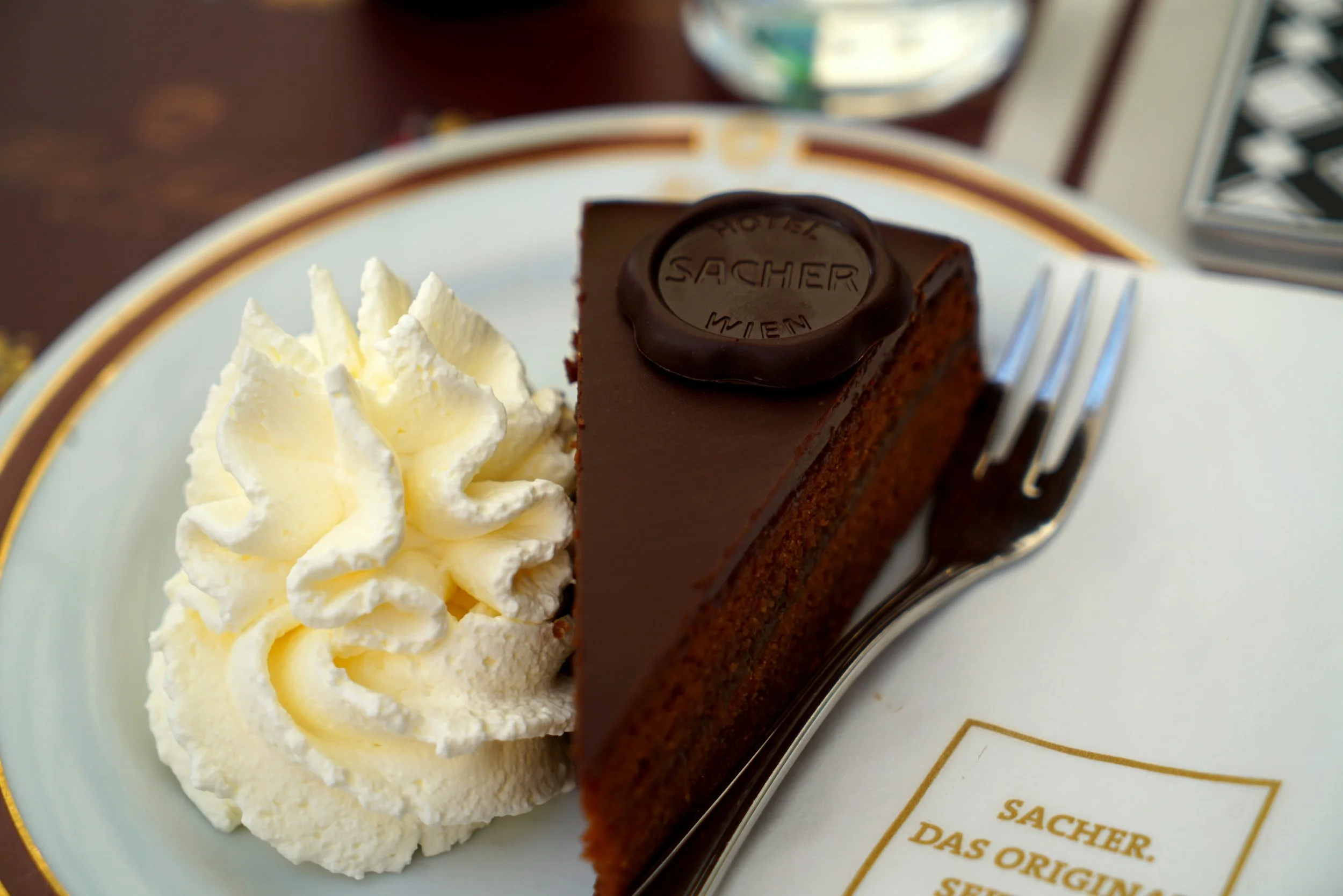 Slice of Sachertorte with whipped cream on a plate, accompanied by a fork.