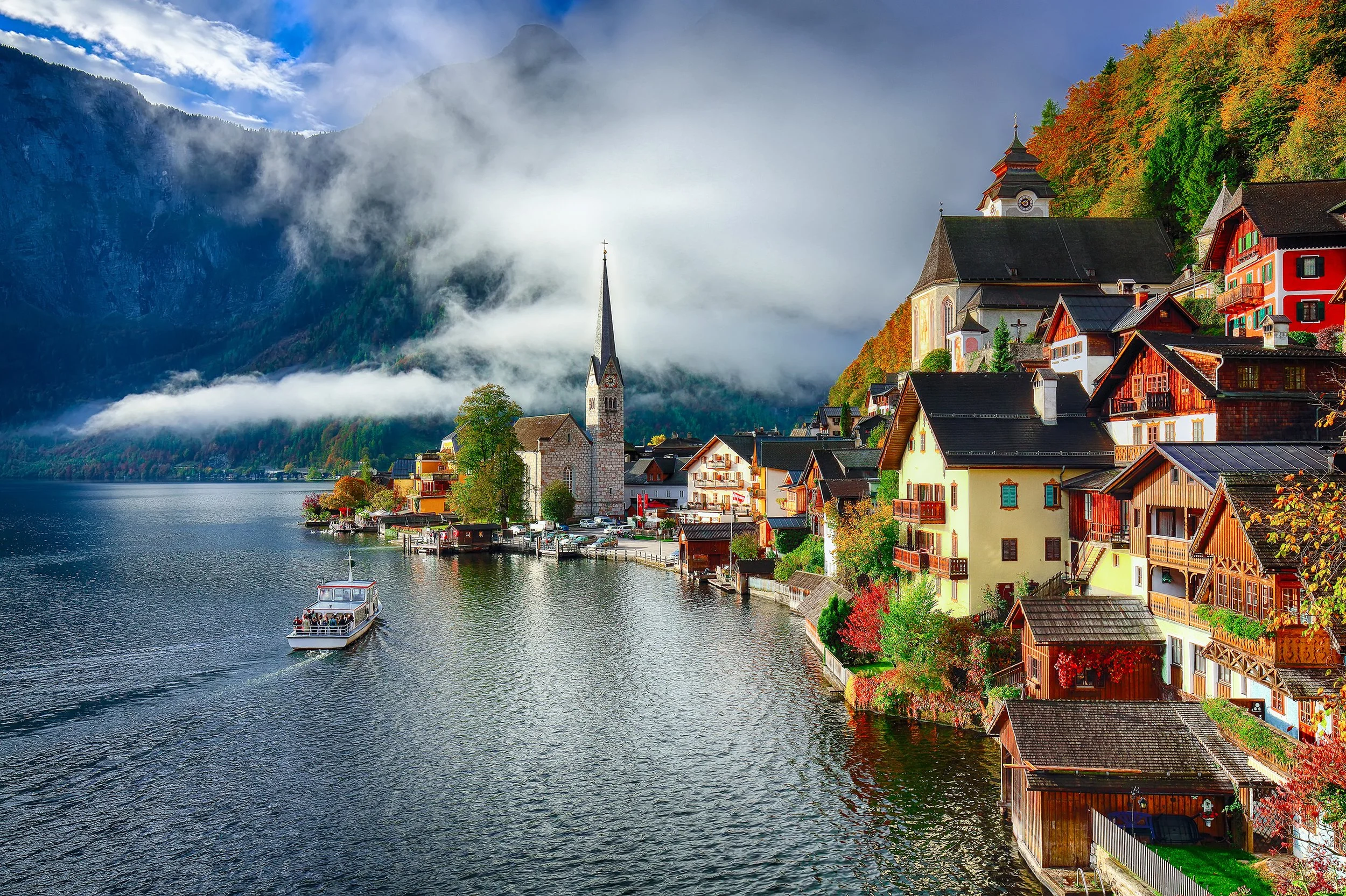 Scenic view of a lakeside village with colorful houses, a church with a tall spire, and misty mountains in the background. A boat is sailing on the water.