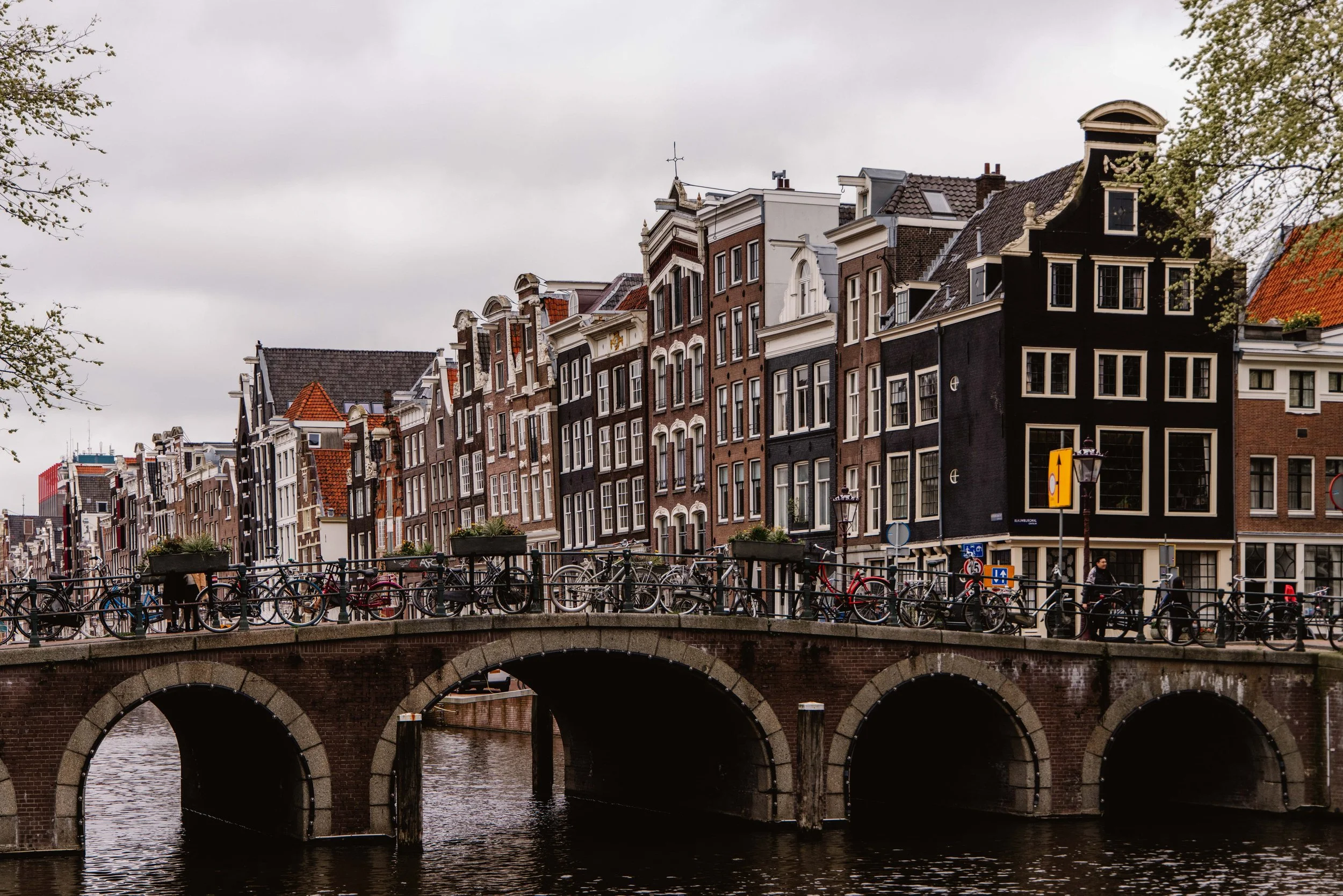 A canal with a stone bridge over it, tightly packed historic buildings with gabled roofs lining the canal, bicycles parked along the railing, and cloudy sky overhead in Amsterdam.