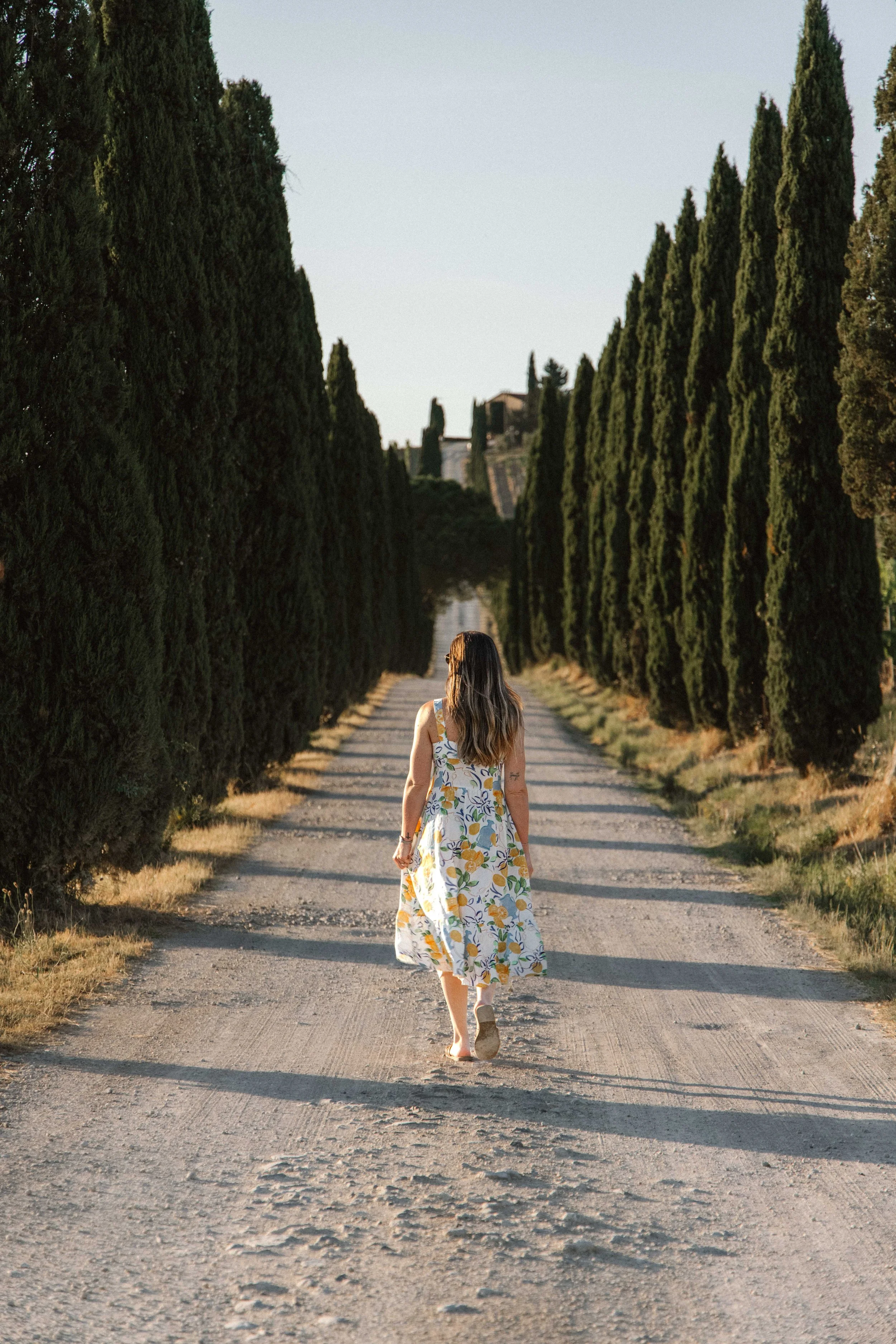 A woman in a floral dress walking down a dirt road flanked by tall cypress trees during daylight.