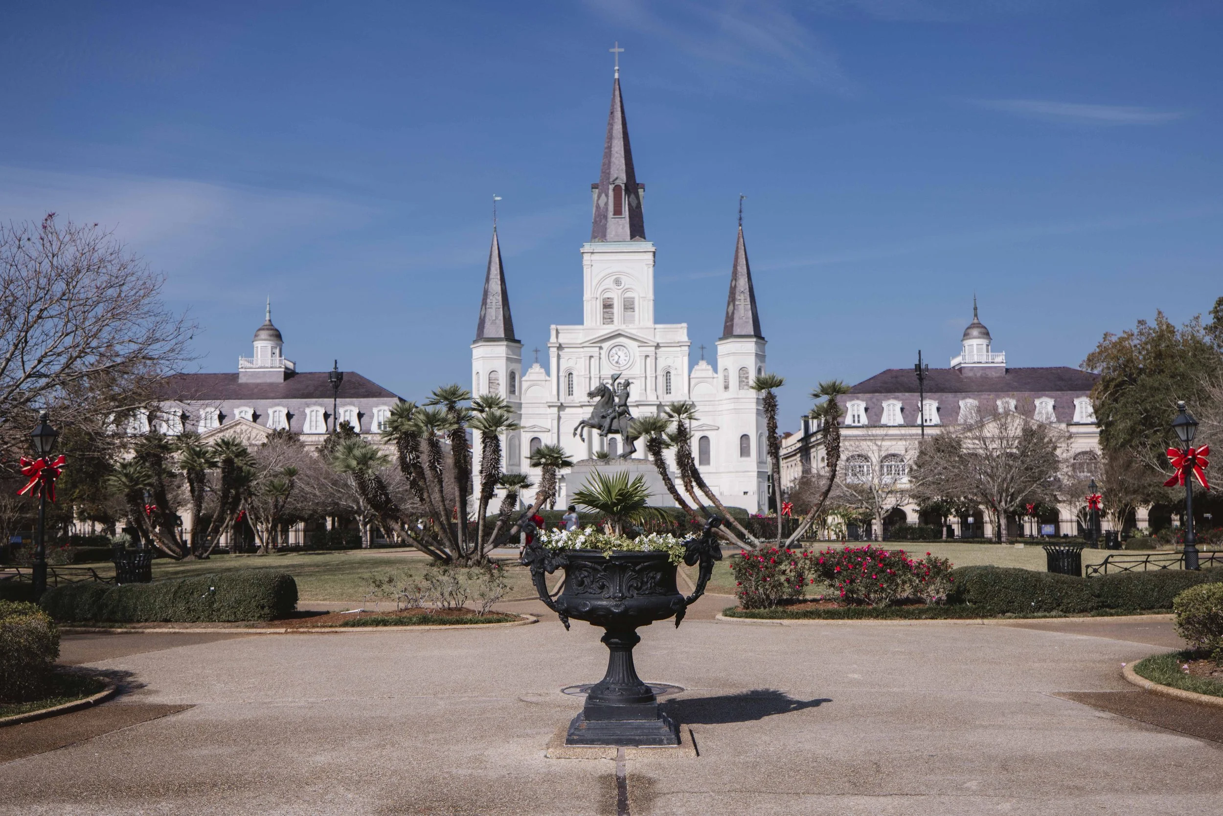 The image shows a large white church with three tall spires against a blue sky. In front of the church, there is a decorative iron planter with a statue of a cowboy on horseback, surrounded by palm trees and landscaped gardens with bushes and flowers. The area is paved with walkways and is decorated for the holiday with red bows on lampposts.