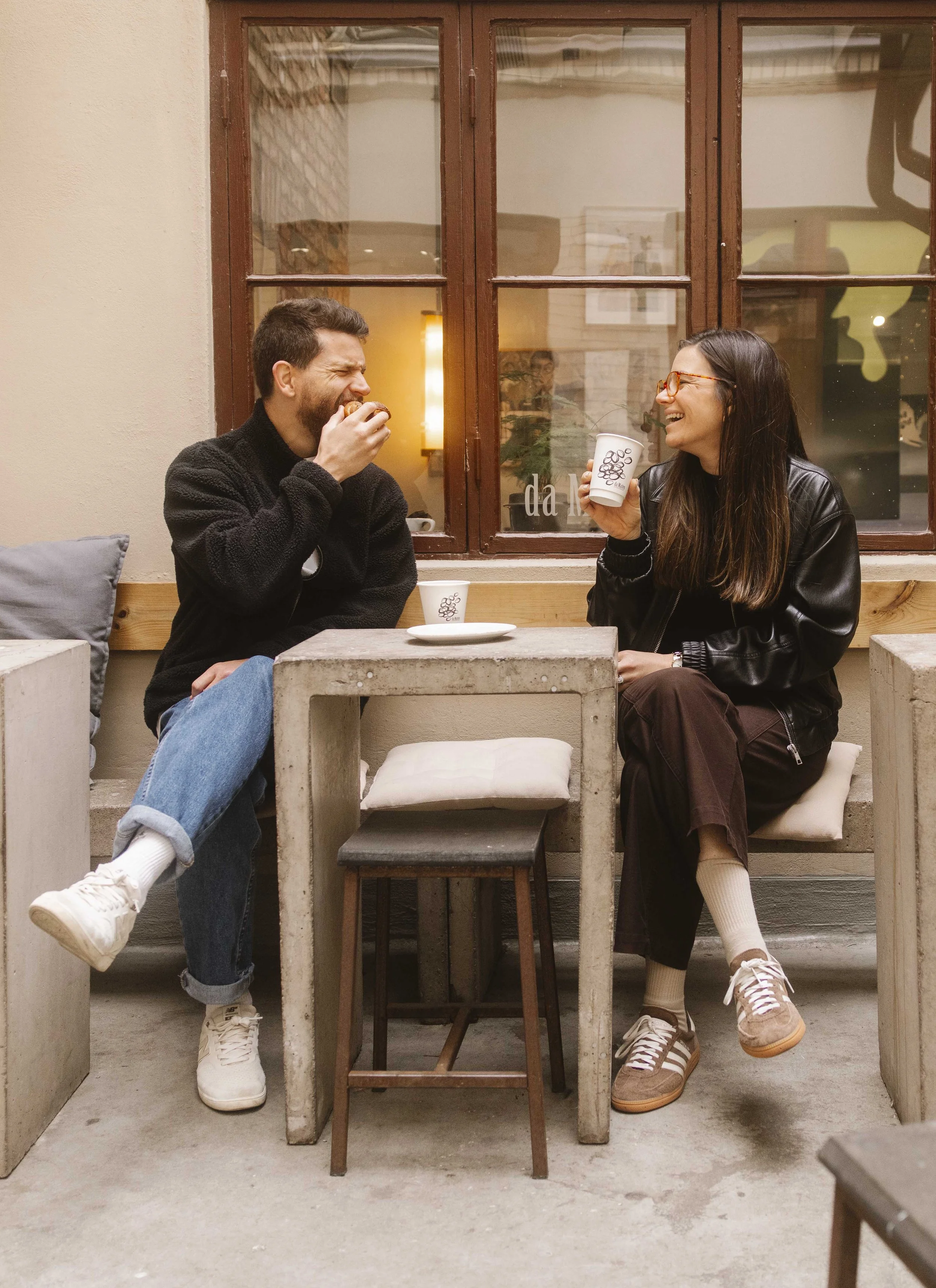 Husband and wife sitting at an outdoor cafe table, laughing and eating. The man is eating a pastry, and the woman is holding a cup, both wearing casual clothing and sneakers.