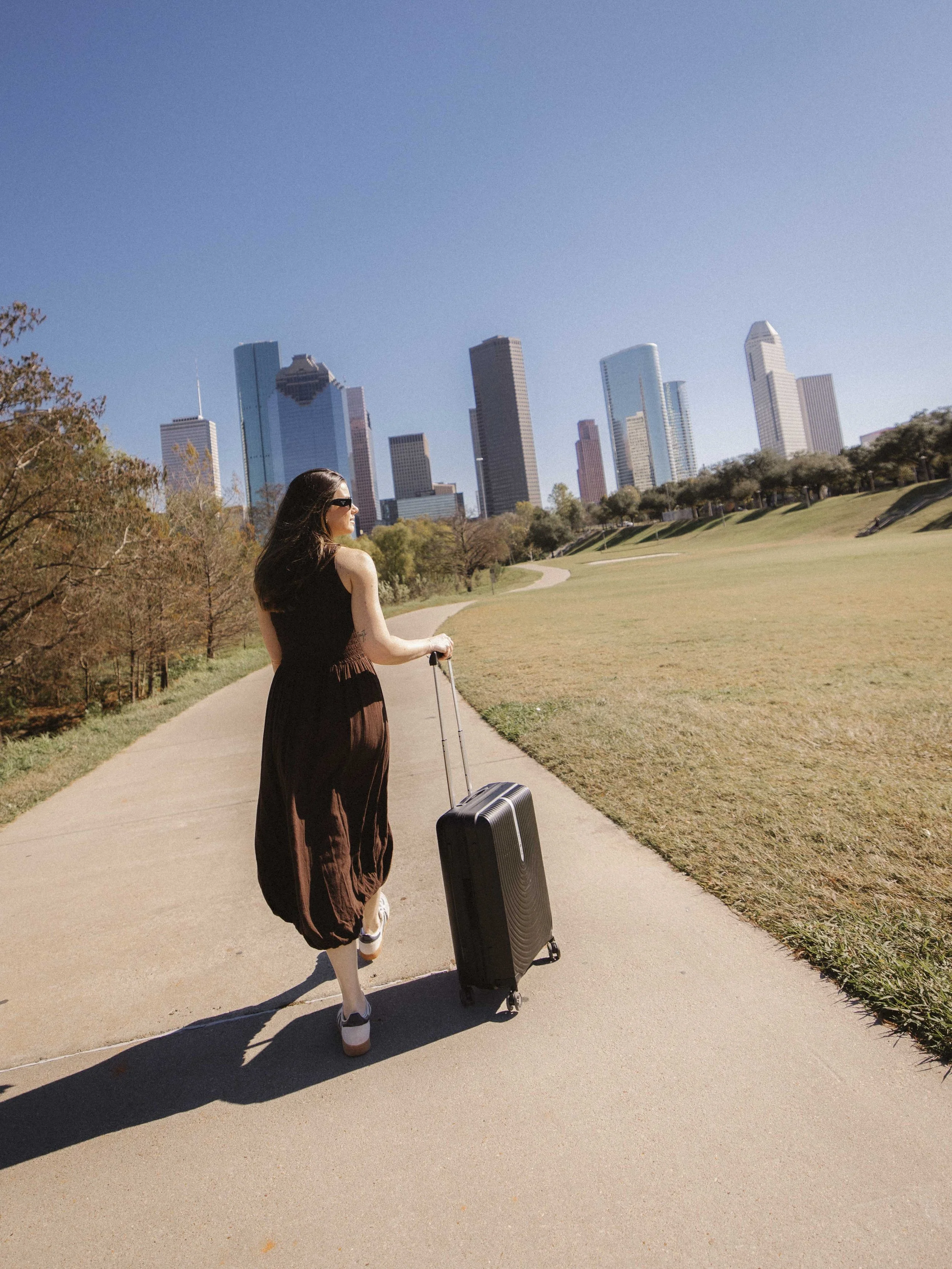 A woman walking along a paved path with a rolling suitcase in a park, with a city skyline featuring tall buildings in the background on a clear, sunny day.