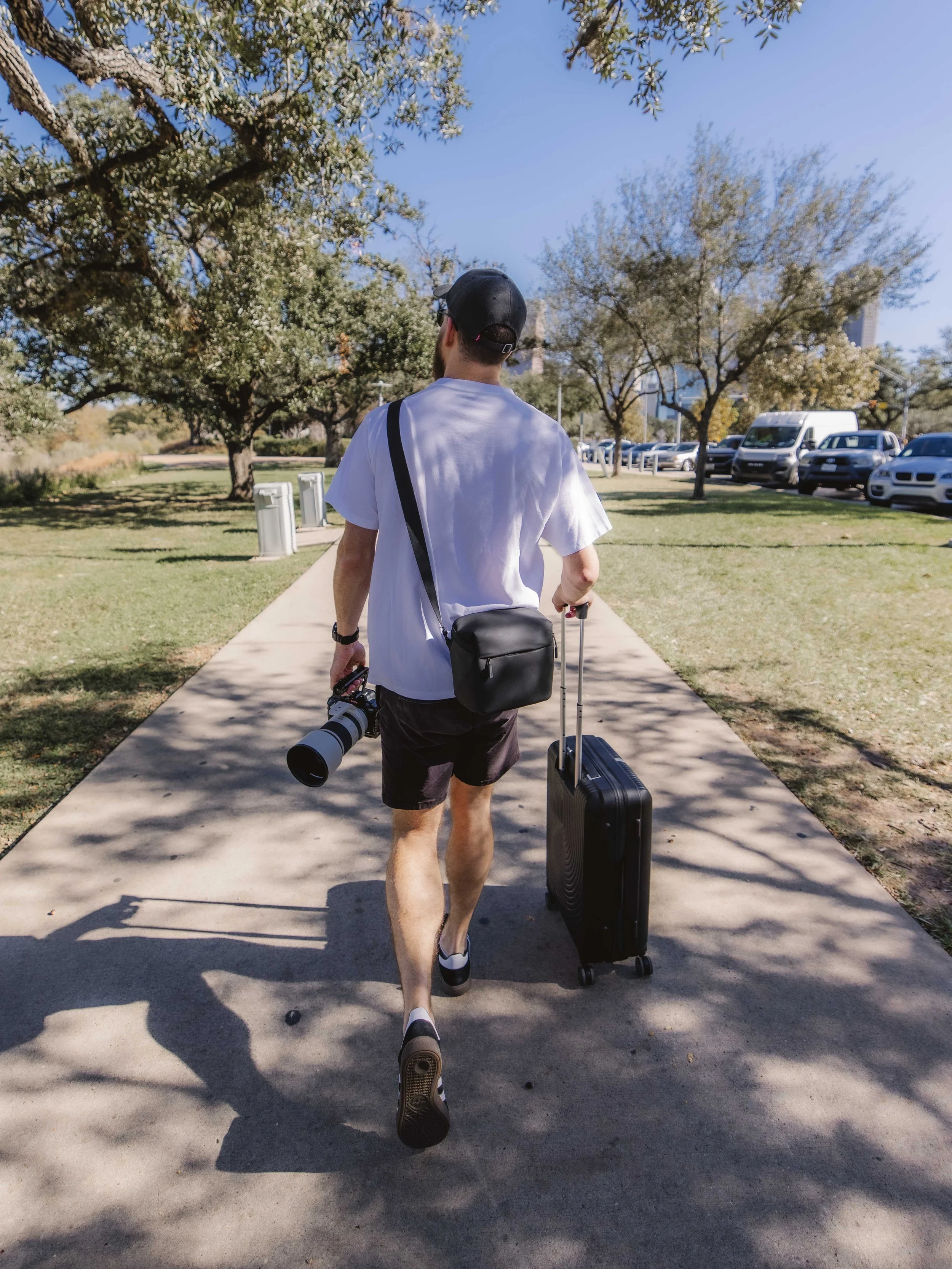 A man walking on a sidewalk with a rolling suitcase and a camera, wearing a white T-shirt, shorts, and a baseball cap, in a park on a sunny day.