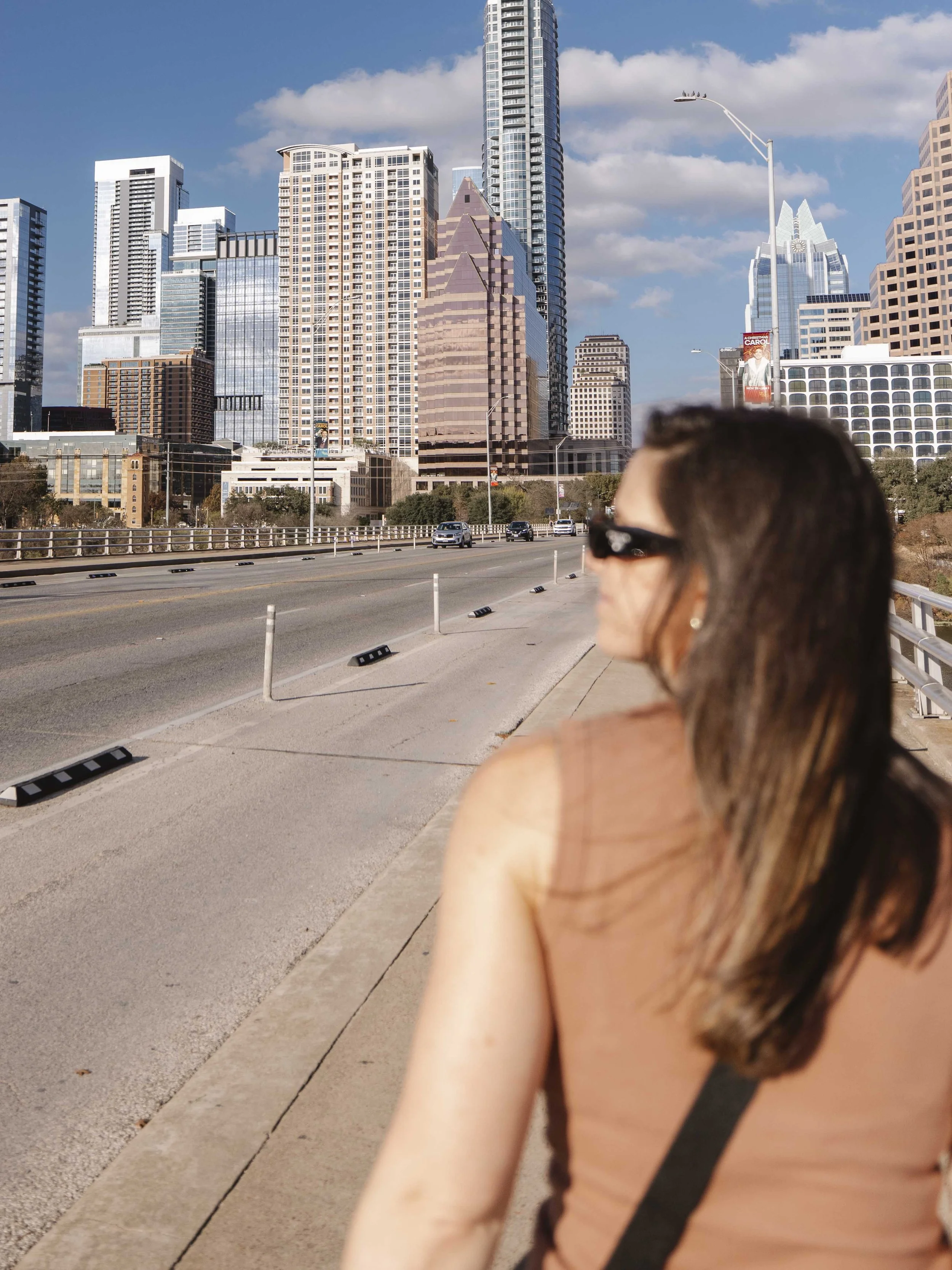 A woman with brown hair wearing sunglasses and a brown tank top, walking along a city sidewalk with a view of tall skyscrapers in the background on a sunny day.