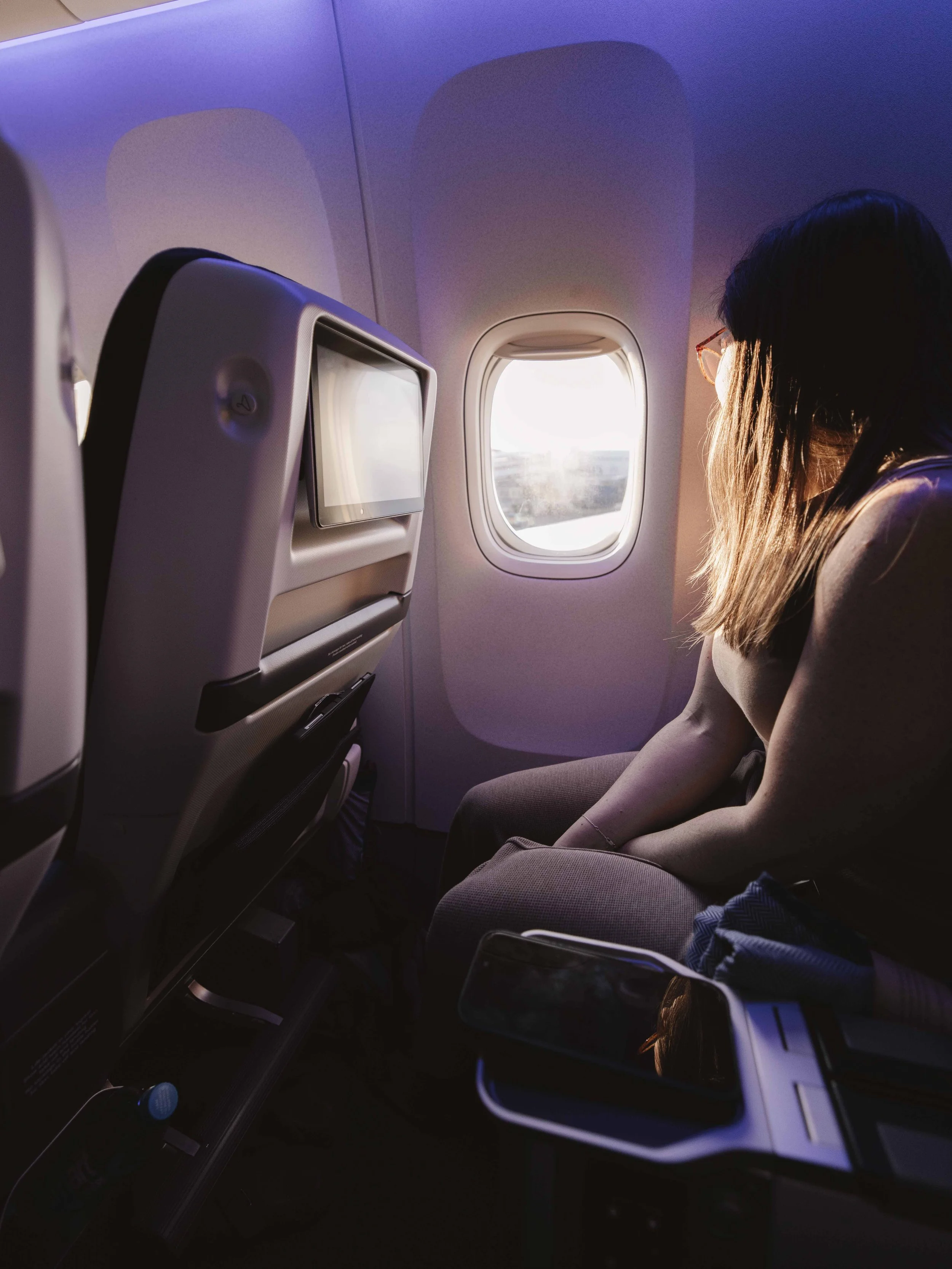 An airplane window seat with a female passenger facing the window, watching the sunset or sunrise.